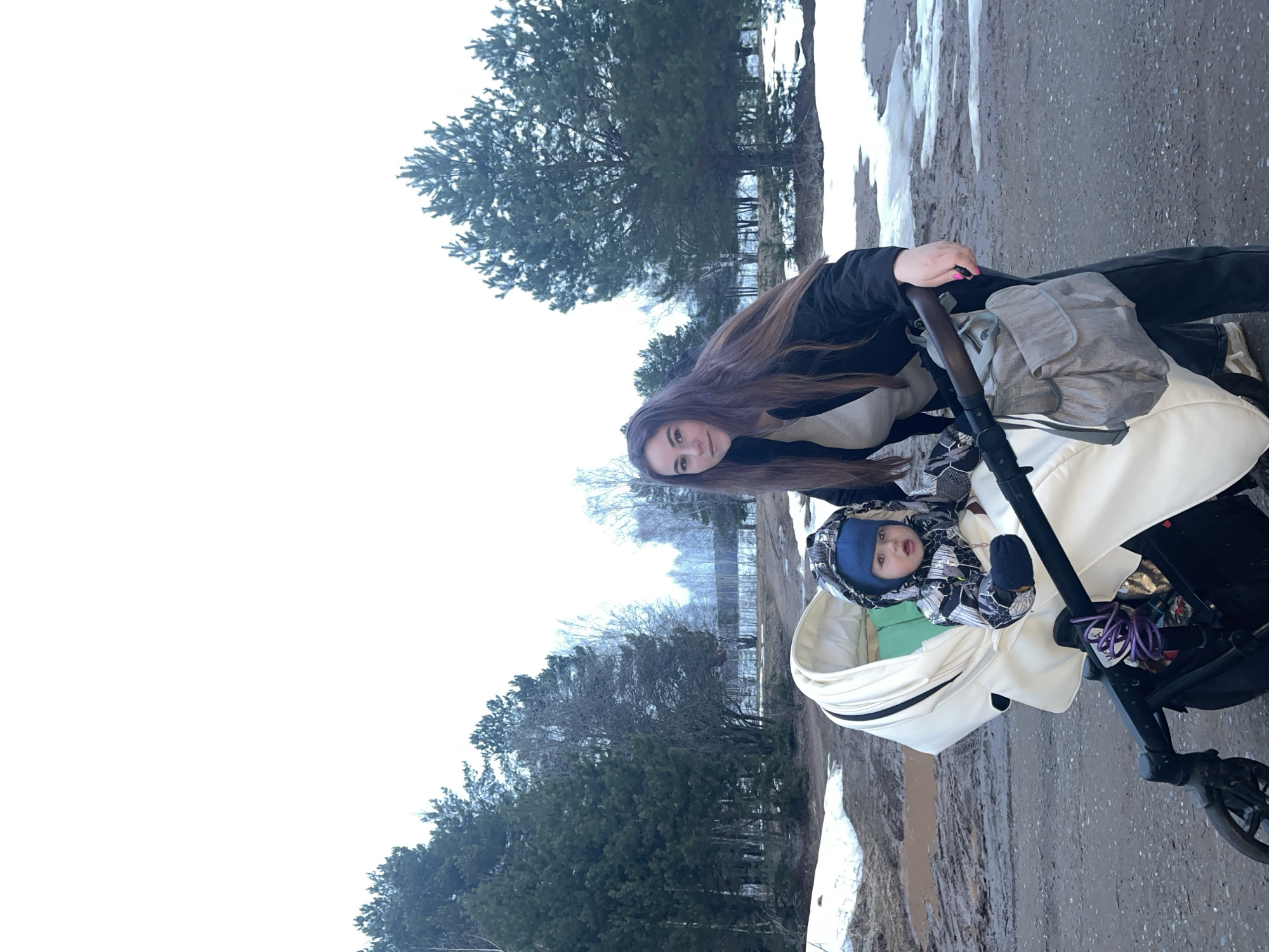 A woman stands beside a baby in a stroller in Chaikovsky, Russia. The stroller is positioned on a muddy path. Trees are visible in the background. There are patches of snow on the ground.