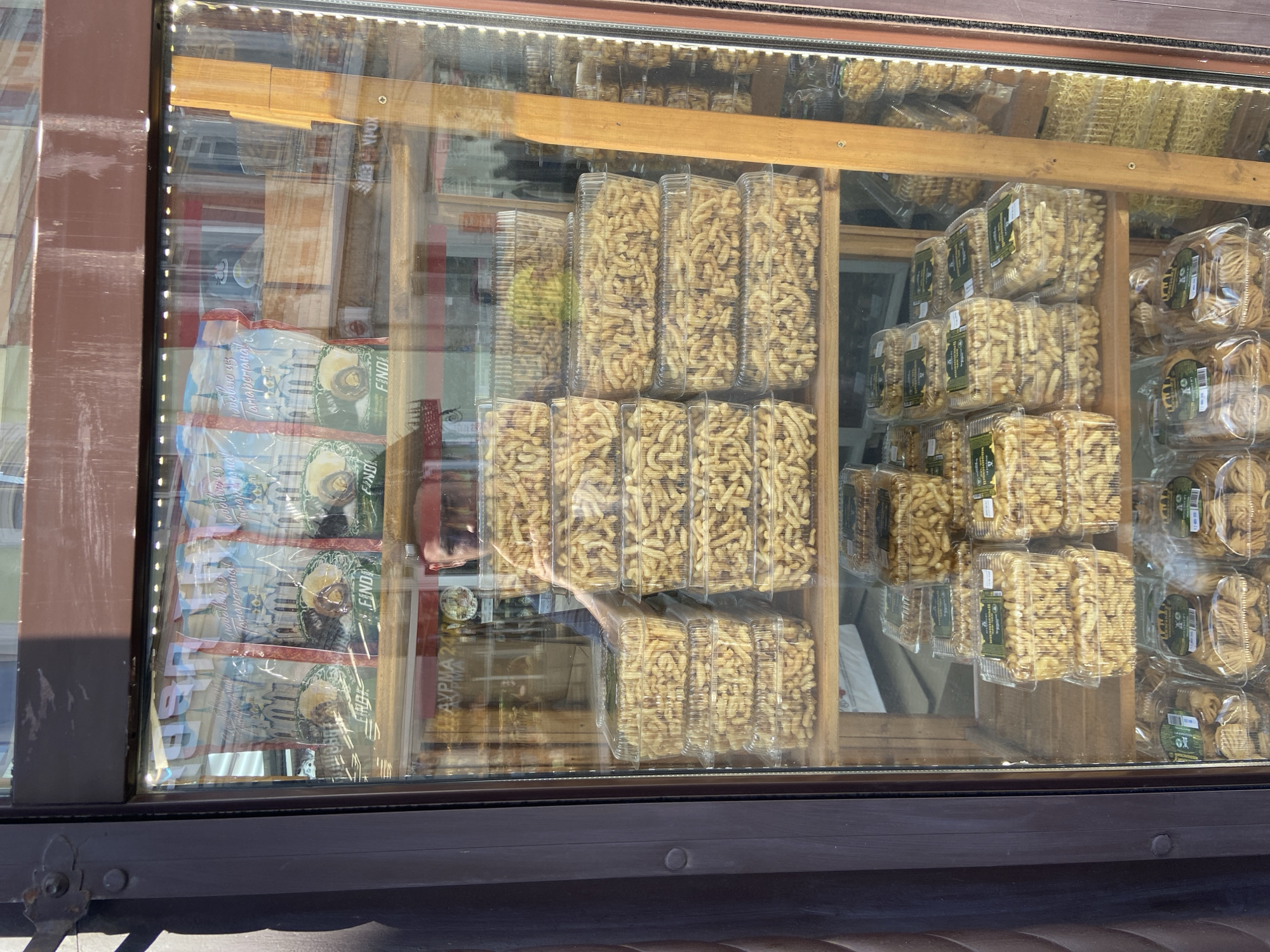 A glass-fronted display cabinet showcases stacks of clear plastic containers filled with a light brown, irregularly shaped pastry or snack. The cabinet is framed by dark brown wood. The pastries are a consistent light brown color and appear to be twisted or looped. They are packaged in rectangular containers, and these containers are stacked neatly on wooden shelves within the cabinet. In the background, reflections on the glass reveal the interior of a store, including a person's face visible in the upper portion of the display. The lower shelves also hold more containers of the same pastry, as well as some that appear to be different shapes or colors. A sign is visible through the glass in the upper section, with Russian text. The overall impression is of a food vendor or shop selling packaged snacks.