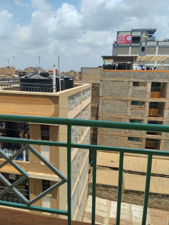 The image presents an elevated view of a densely built urban environment within Witeithie ward, Kenya, under a partly cloudy sky. In the immediate foreground, a green metal railing with a diamond pattern is visible, suggesting a balcony perspective. The midground features multiple multi-story buildings constructed primarily from exposed concrete blocks and some finished with light-colored paint. Several rooftops are equipped with large black cylindrical water storage tanks. On one rooftop to the right, clothes are hanging on a line to dry. The lower areas between buildings reveal corrugated metal roofs of smaller structures. In the background, a taller, multi-story building displays a red and white "TO LET EXECUTIVE APARTMENTS" sign with contact numbers: 0700 22 44 33 and 0711 22 44 33. Distant structures and communication masts are visible on the horizon. No individuals or active interactions are depicted.