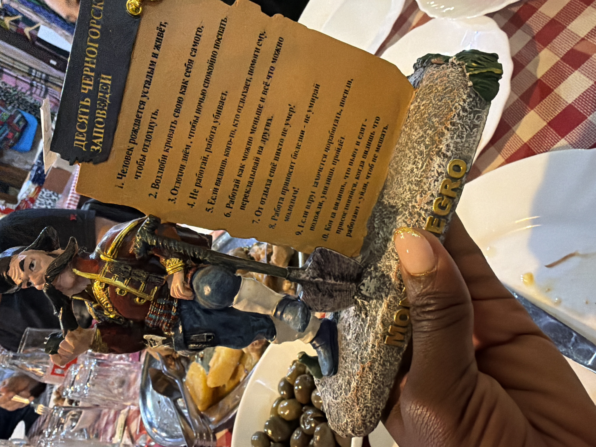 A hand holds a souvenir statuette from Montenegro, inscribed with “Черногорск” in Cyrillic. The figurine depicts a man with a shovel climbing a rocky formation. Attached to the statue is a paper with "ДЕСЯТЬ ЧЕРНОГОРСК ЗАПОВЕДЕЙ" ("Ten Montenegrin Commandments") written in Cyrillic. The commandments humorously relate to avoiding work and prioritizing rest. In the background is a table set with plates and dishes, with other diners visible.
