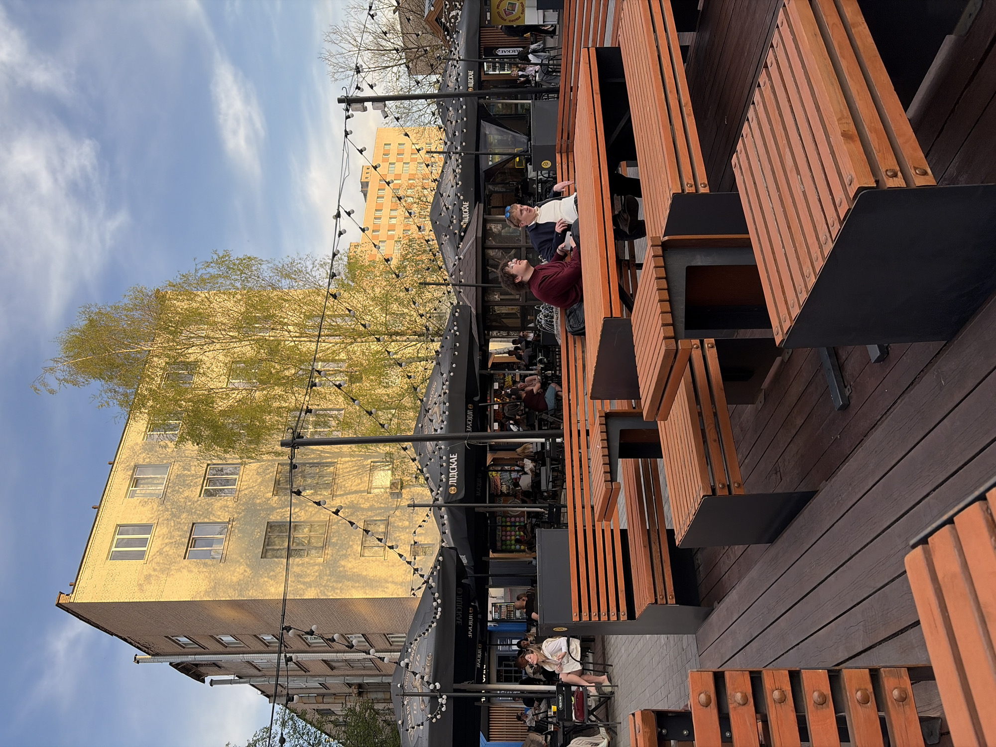 The image depicts an outdoor seating area in Minsk, Belarus. Several people are seated at wooden tables with black accents. String lights hang above the tables. Buildings are visible in the background, including one with a yellow brick facade. Trees with green foliage are also present. Patrons appear to be eating and socializing. A black awning with the word "ПІДСАСКЕ" (PIODSATSKE) printed on it is visible above one of the seating areas.