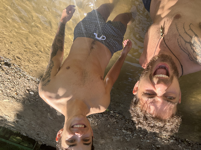 Two males are depicted waist-up, standing in shallow, murky water in Ban Mai Saha Sampan, Thailand. The male on the left, positioned closer to the camera, has curly brown hair, a beard, and a goatee. He exhibits an open-mouthed expression with bared teeth, eyes narrowed, and wears a dark necklace with a light-colored pendant. Tattoos are visible on his left shoulder and arm. The male on the right, positioned slightly behind and to the right, has short dark hair and is smiling broadly. He has intricate tattoos on both arms, including what appears to be a wolf on his left bicep. He wears dark swim trunks with a white patterned design and a white drawstring. Both individuals are shirtless. The water, a light brown hue, covers their lower legs. To the right and behind the subjects, the environment consists of a light brown, rocky, and earthy bank, suggesting a natural stream or river setting under daylight conditions.