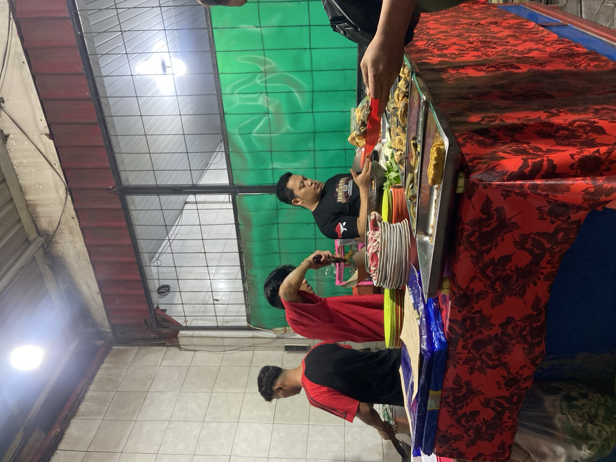 This image depicts an outdoor food stall in Balonggabus, Indonesia. The scene is illuminated by artificial lighting. In the foreground, a table is covered with a red tablecloth featuring a dark floral pattern. On the table, there are stacks of plates, some green and yellow, and trays filled with prepared food items. Several people are visible behind the table. On the left, a person wearing a red and black shirt is standing and appears to be cooking. In the center, another person in a red shirt is actively preparing food, holding utensils. To the right, a person in a black t-shirt with text and an emblem is looking at food on a red plate. Another individual's hands are visible on the far right, holding a red plate. The background shows a green tarp or curtain with a white pattern, and part of the stall's structure including metal beams and what appears to be corrugated metal roofing. The overall environment suggests a busy, informal food service setting at dusk or night.