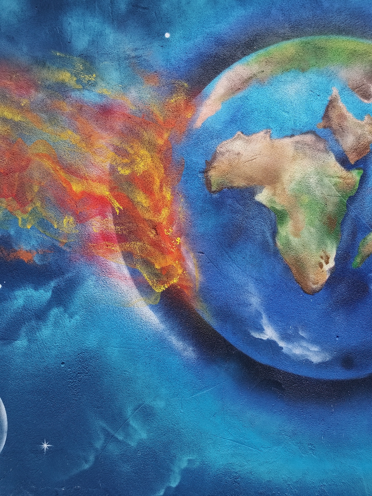 A close-up image depicts a mural featuring an Earth-like planet with Africa prominently visible. To the left of the planet, vibrant red, orange, and yellow flames erupt outwards. The background is a textured blue, suggesting space, with several stars and a partial moon visible. The location is Unknown, Unknown.