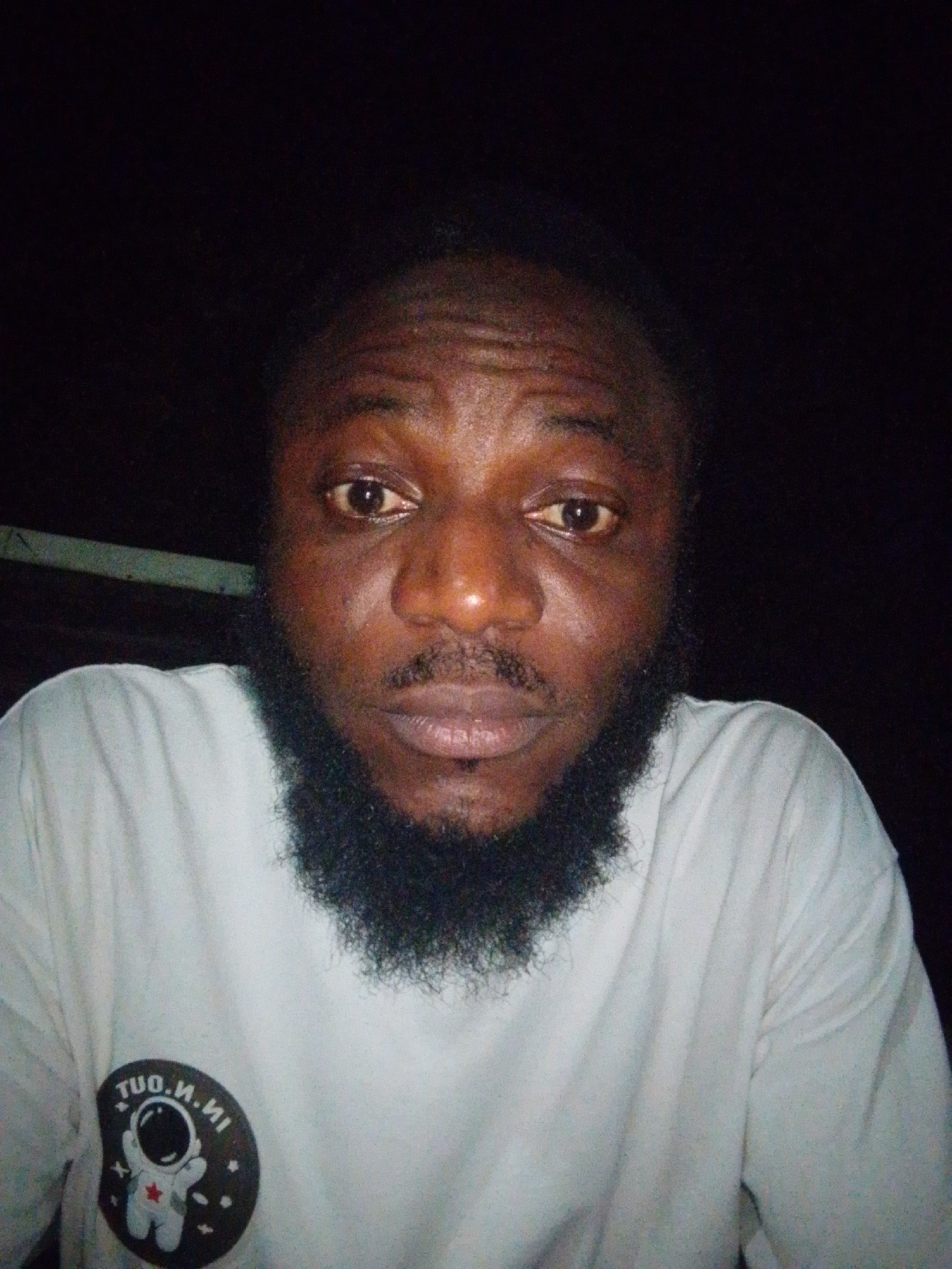 A close-up portrait, likely a selfie, captures a man with dark skin, a full black beard, and dark hair. He is looking directly at the camera with a neutral to slightly solemn expression. He wears a light-colored, possibly white, t-shirt. The background is uniformly dark, indicating the photo was taken at night or in a very dimly lit setting. The image exhibits a noticeable grain, typical of low-light photography. On the left side of his t-shirt, there is a circular patch featuring a white astronaut illustration. The text around the astronaut is mirrored, reading "TUO.N.NI," which would be "IN.N.OUT" if unmirrored. No specific location cues are visible within the image, but the provided context places the scene in Lokoja, Nigeria.