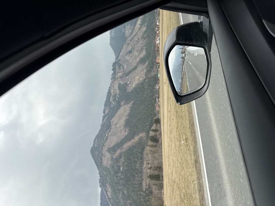 The image depicts a panoramic view from inside a moving vehicle on a road in Teufenbach-Katsch, Austria, under an overcast sky. The foreground on the right shows the dark gray interior trim of a car door, and a black side-view mirror is prominent in the lower right, reflecting the road behind. The primary scene outside the window features a two-lane asphalt road with a white shoulder line and a broken white center line, running alongside a wide, dry, light brown field. Beyond the field, a cluster of residential buildings with varied roof colors, including red and orange, forms a village. In the background, large mountains covered in a mix of coniferous forest and areas of lighter, possibly deforested or sparsely vegetated slopes, rise under a cloudy, grey sky. A faint white aircraft contrail is visible diagonally across the upper-left sky. The side-view mirror reflection shows the road extending behind the vehicle, with additional cars, buildings, and a distinct church spire suggesting a concentrated settlement, consistent with a view approaching or leaving a village like Teufenbach within the Teufenbach-Katsch municipality.