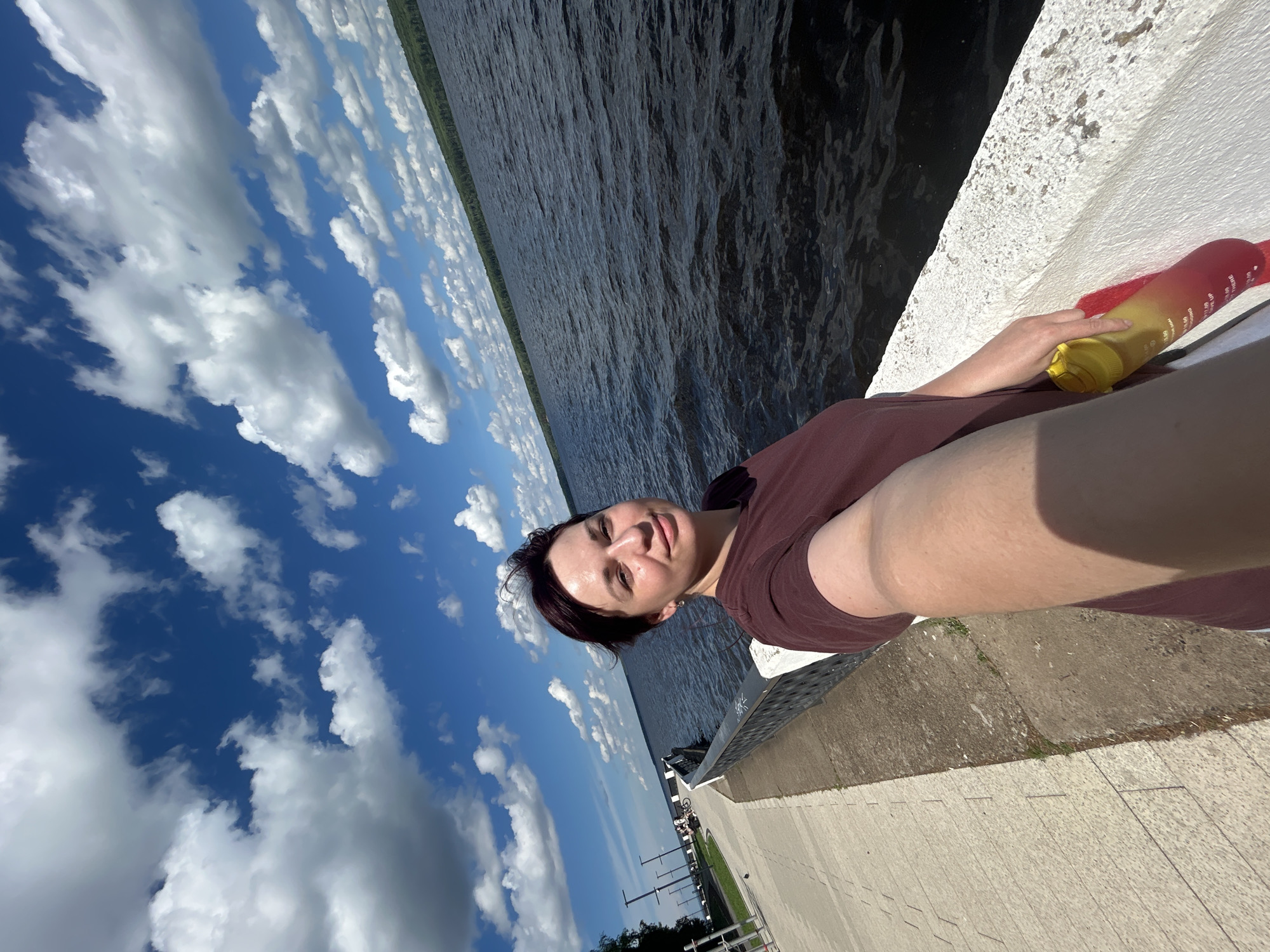 A woman with dark hair stands on a paved walkway next to a body of water, holding a large water bottle. She is taking a selfie, smiling at the camera. The walkway is bordered by a railing, and a body of water stretches out before her. In the background, a treeline is visible, and the sky is filled with white clouds against a bright blue background. The scene appears to be a waterfront promenade in the city of Chaikovsky, Russia.