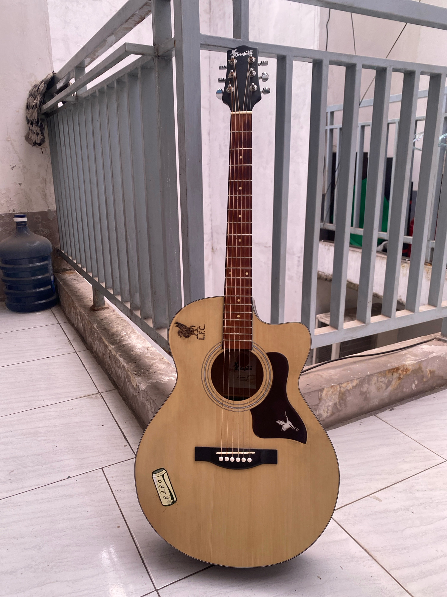 An acoustic guitar, branded "Kapferg," with a light wood finish and black pickguard, stands upright on a tiled floor. The guitar features a cutaway body, six strings, and a headstock with tuning pegs. A sticker depicting a can with the text "C.R.O.O." is affixed to the lower bout of the guitar. To the left of the guitar, a large blue water jug rests on a concrete ledge. Behind the guitar is a grey metal railing, part of a balcony or fence, with a piece of patterned fabric hanging from it. The setting appears to be an outdoor area with a light-colored tiled floor and a concrete wall. This scene is located in the city of Semarang, Indonesia.