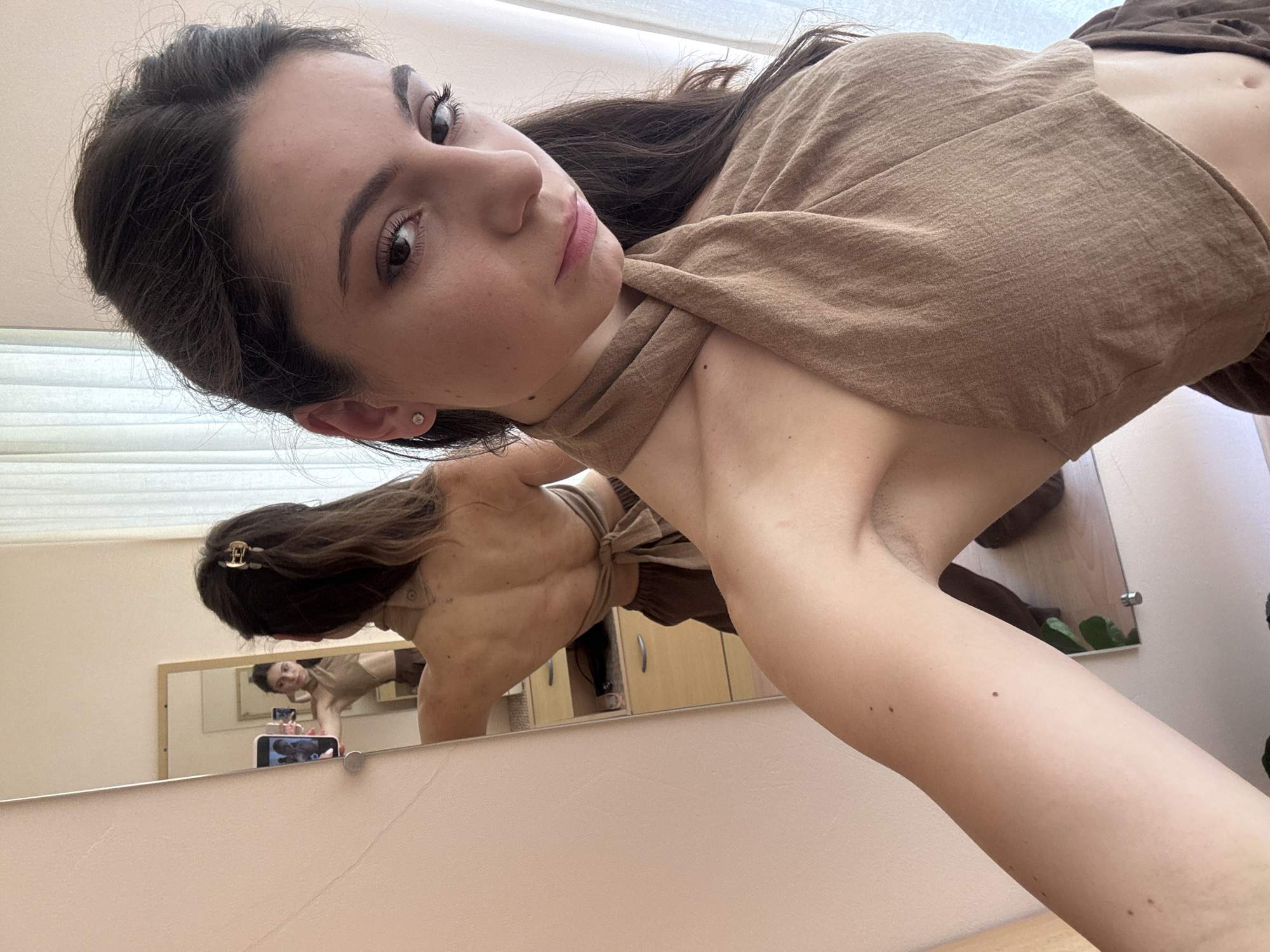 In a dimly lit room, a young woman with dark hair tied back in a ponytail takes a selfie in a mirror. She is wearing a brown halter top and brown pants. Her back is visible in the reflection, showing a toned physique. The room appears to be a bedroom or dressing area, with a dresser and closet visible in the background. The overall impression is one of a casual, personal moment captured in an everyday setting.