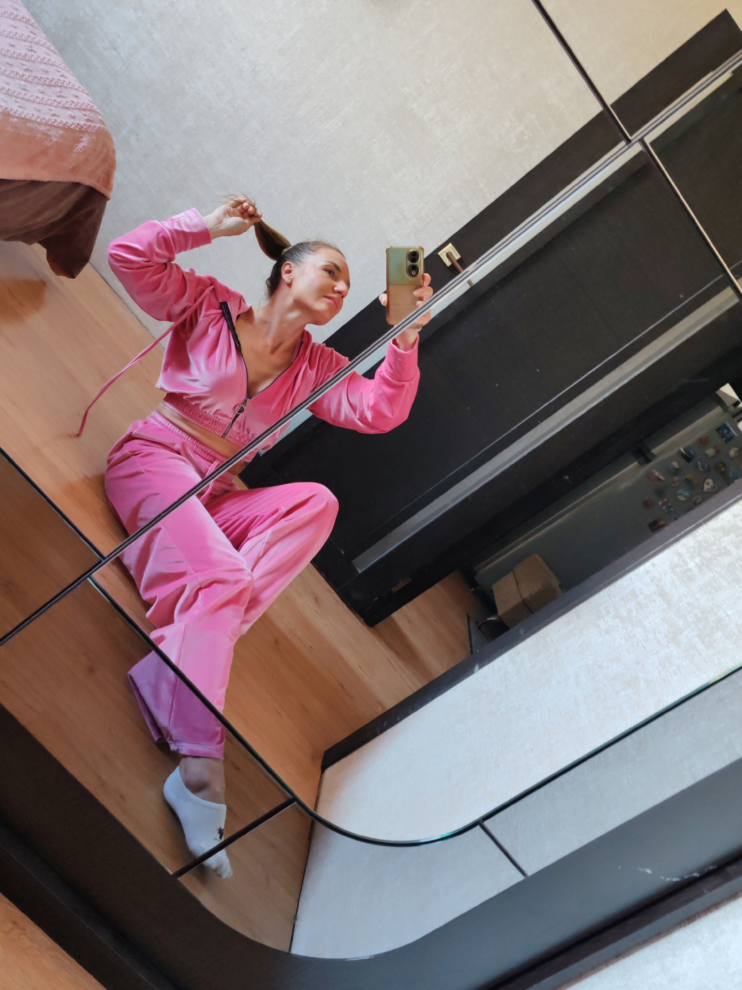 An adult female, positioned on a segmented mirror surface, captures a selfie using a smartphone held in her right hand. She has light skin, brown hair styled in a high ponytail, which her left hand grasps. Dressed in a bright pink velvet cropped zip-up tracksuit and white ankle socks, she presents a subtle smile or smirk while looking towards the phone's reflection. The mirror's dark framing lines dissect the scene, reflecting a light wooden floor beneath her. Background reflections reveal a bed with a pink textured blanket on the left, and a dark wardrobe or wall unit with thin silver accents on the right. Further into the reflected background, a light-textured wall and a refrigerator adorned with numerous magnets are visible. The scene takes place indoors, within what appears to be a bedroom or dressing area in Petropavlovsk-Kamchatsky, Russia.