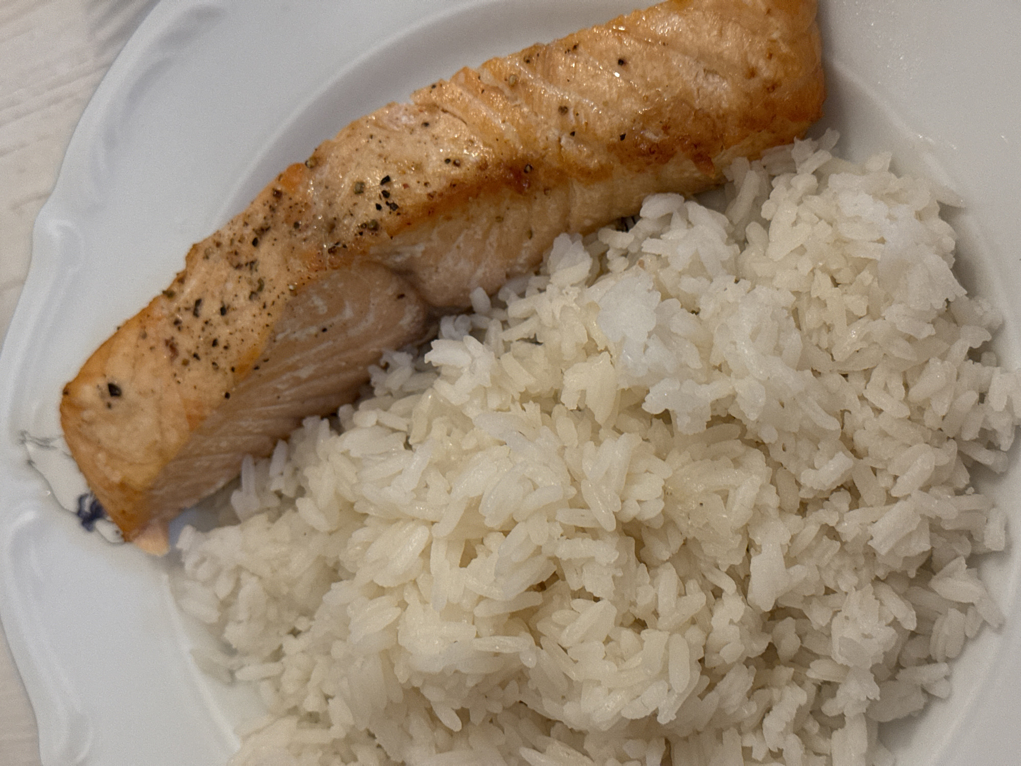 A white plate holds a piece of cooked salmon and a pile of white rice. The salmon has a browned top with visible seasonings. The rice is fluffy and slightly sticky in appearance. No people are visible. The scene does not appear to have any characteristics specific to Chaikovsky, Russia. The setting is a neutral, possibly indoor, environment.