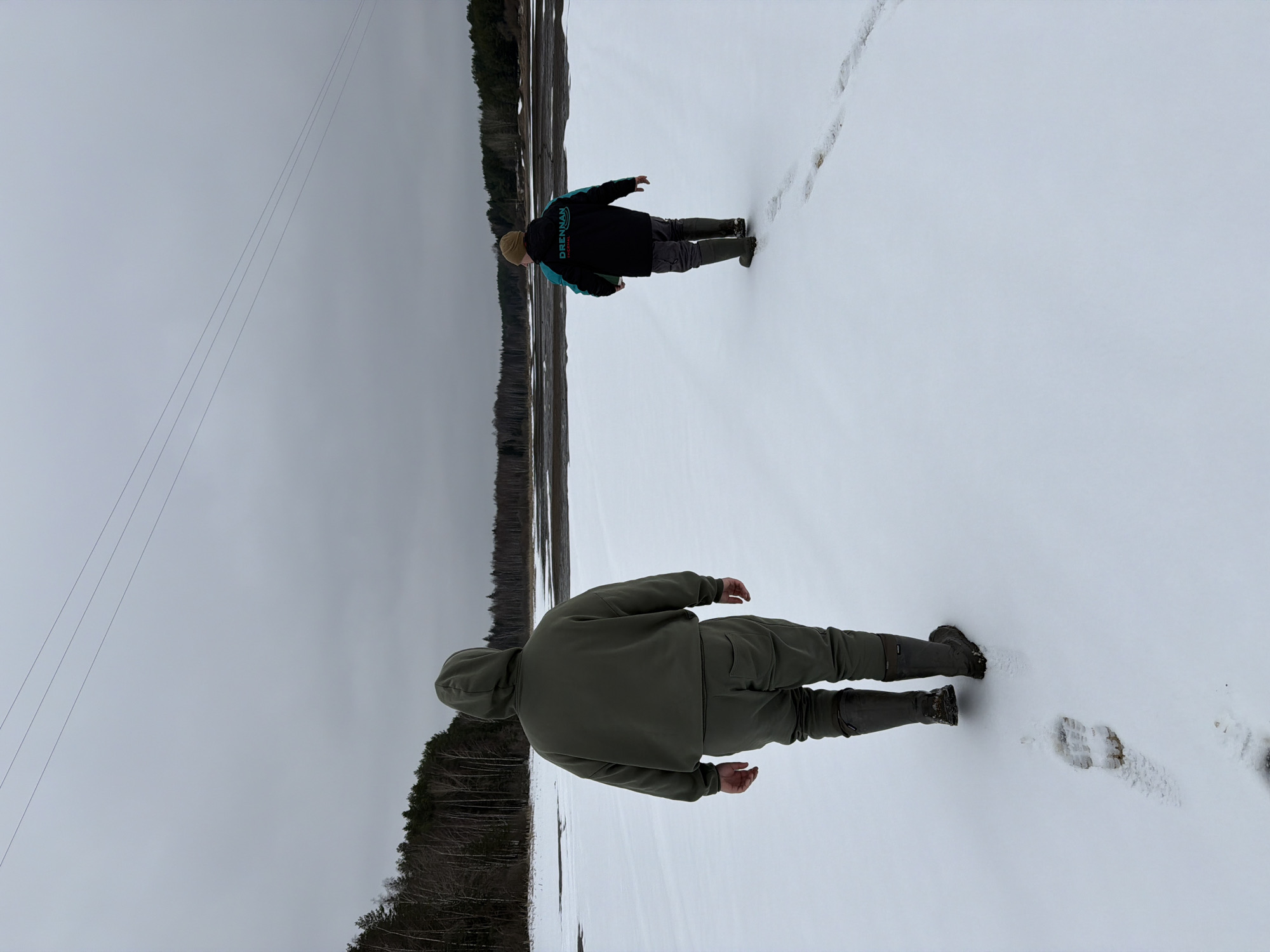 The image depicts a snowy, flat landscape with two individuals walking across it. The environment appears to be a field or frozen lake, bordered by a line of trees in the background. The sky is overcast.
One individual is wearing a dark jacket with a turquoise logo, gray trousers, and rubber boots. The second individual, closer to the foreground, is wearing a green hooded outfit and rubber boots. Both individuals are walking away from the viewer.
There are footprints in the snow suggesting a path has been taken. Overhead power lines are visible in the sky.
Due to a lack of specific landmarks in the image, the exact location in Chaykovsky, Russia cannot be definitively determined. The scene suggests a rural or undeveloped area on the outskirts of the city.