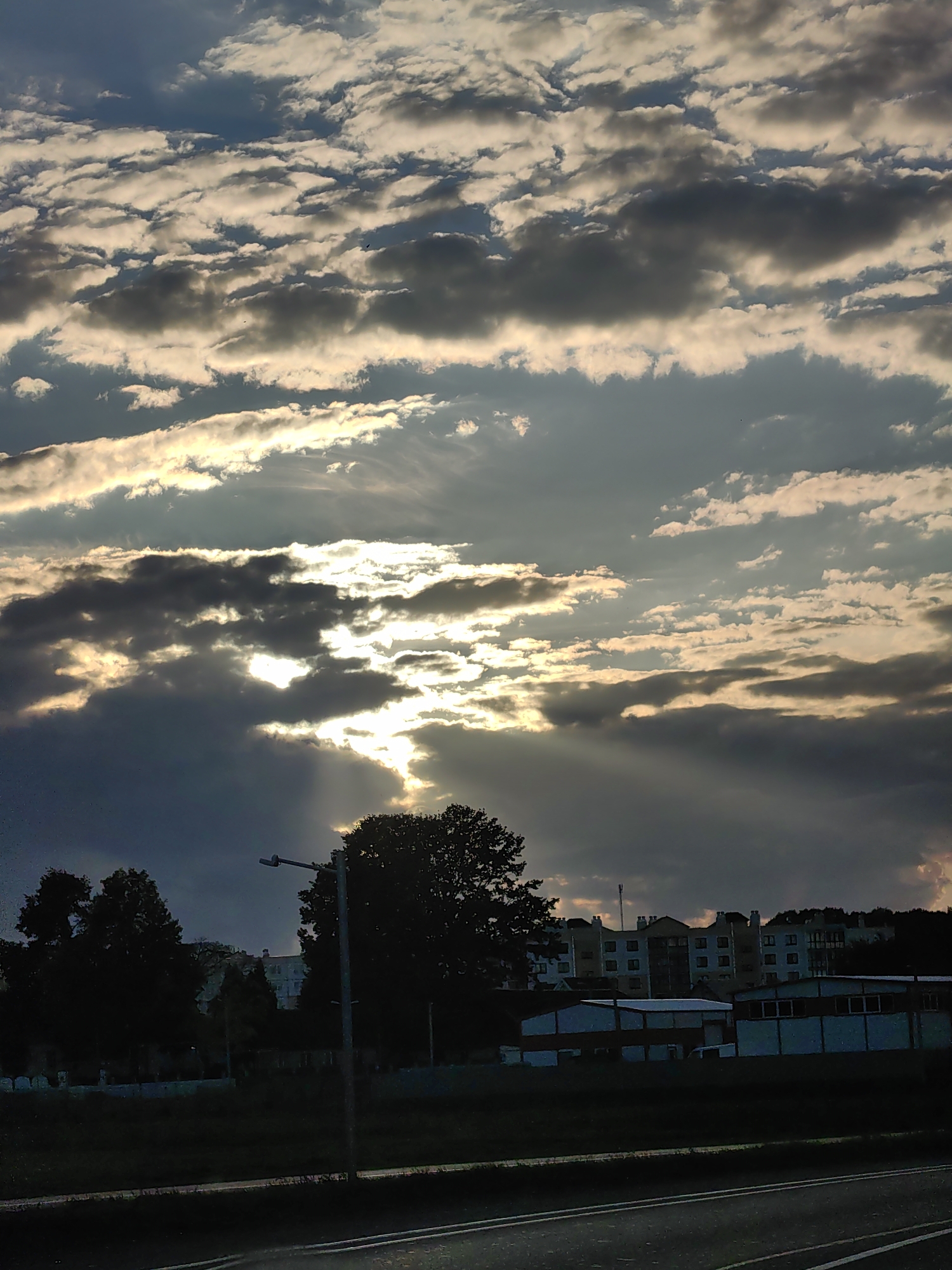 The image captures a cloudy sky over Dzyarzhynsk, Belarus, likely during a sunset or sunrise. The sun is partially obscured by dark, heavy clouds, allowing shafts of light to pierce through, creating a dramatic effect. Below the sky, silhouettes of trees and a lamppost are visible in the foreground. Further in the background, a row of apartment buildings is seen on a slight incline, with smaller industrial-style buildings in front of them. The overall scene suggests an urban or suburban environment at dusk or dawn.