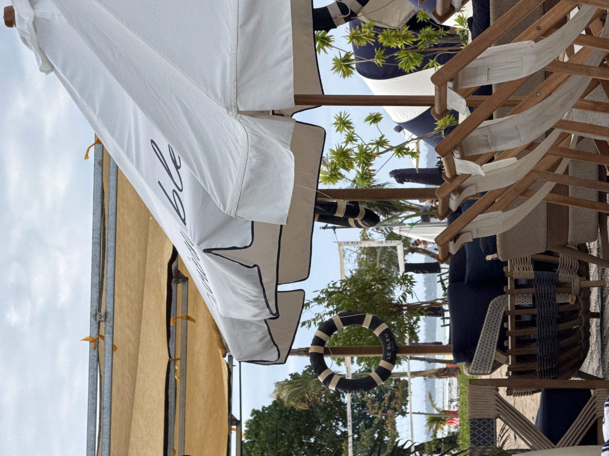 The image depicts an outdoor scene in Kuta Selatan, Indonesia, likely at a beachside venue. Dominating the view are beach umbrellas, some with the word "bleu" written on them. The umbrellas offer shade over beach chairs stacked neatly, indicating a setup for relaxation and leisure. Additionally, a black and white striped life preserver bearing the name "LE BLEU" is noticeable. The environment includes greenery with trees and palm trees, suggesting a tropical setting. In the background, there's a glimpse of the ocean and a sandy beach, with people visible in the distance. Overall, the scene captures the atmosphere of a seaside resort or club.
