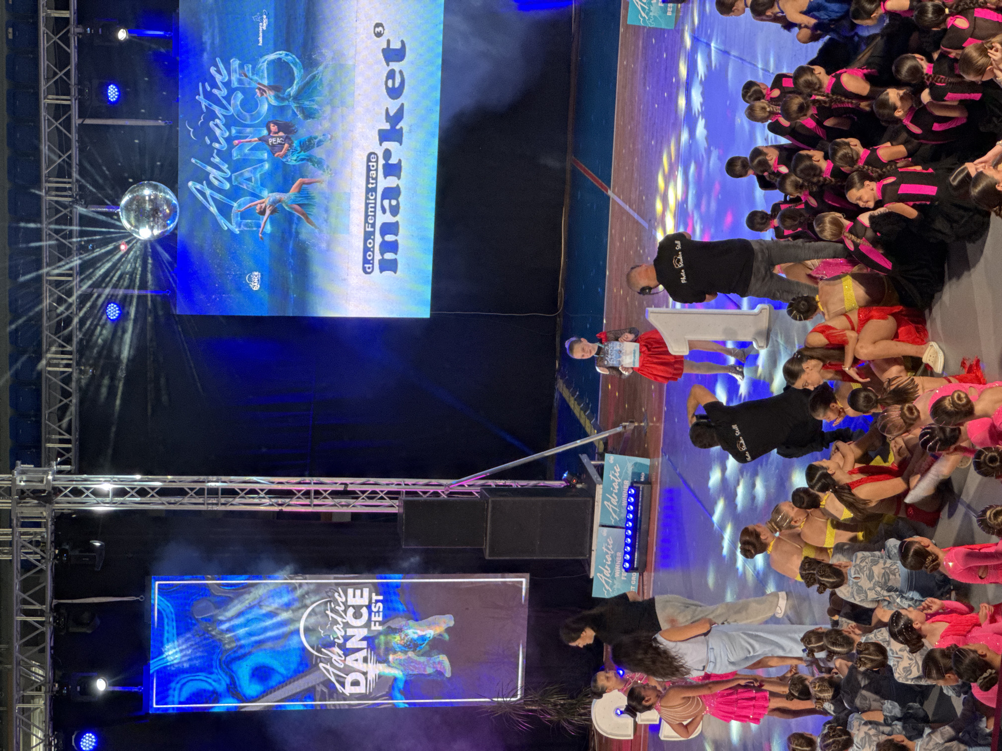 The image depicts a dance competition event at an indoor venue, likely a sports hall, in Bar, Montenegro. On the left side, a large vertical banner displays "Adriatic DANCE FEST" with abstract blue and green imagery. A large screen in the background shows "Adriatic DANCE" and promotional content for "d.o.o. Femic trade market³." A disco ball hangs from the ceiling.
On the stage, several groups of young dancers are present. One young girl in a red dress stands near a large number '1' object, holding a certificate. Several adult women are also visible, some appearing to be instructors or organizers, while others are participants. A photographer, wearing a black t-shirt with "Photo Studio Still" printed on it, is actively taking pictures.
The floor of the venue is illuminated with colorful stage lighting, creating a vibrant and dynamic atmosphere. The majority of the dancers, dressed in various performance costumes including bright pink, yellow, and black outfits, are seated or kneeling on the floor in front of the stage, awaiting their turn or having completed their performances. Some dancers are interacting with each other, while others appear to be resting. The overall scene suggests the culmination of a dance event, possibly involving awards or final performances.