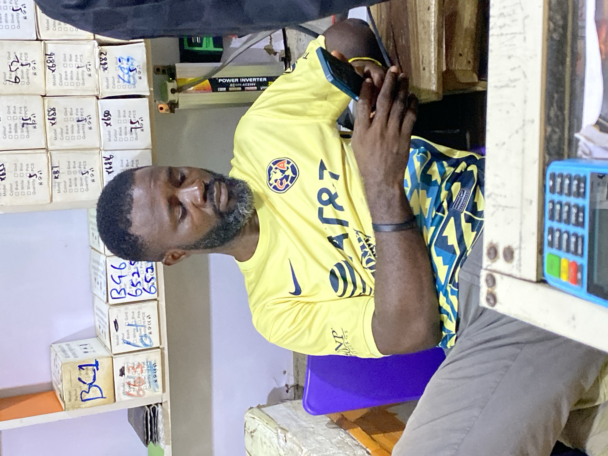 A man is seated in what appears to be a small shop in Lokoja, Nigeria. He is holding a smartphone to his ear, engaged in a conversation. The man has a beard, a yellow shirt with a sports logo, and a black wristband. He's sitting on a purple plastic chair.
The shop's shelves are stocked with small cardboard boxes with handwritten labels, including model numbers "B46", "6525", "652", and "KB1", and color options "Black", "Gold", "White", "Blue", "Silver", and "Pink". Each box indicates a quantity of "10 PCS". A "POWER INVERTER DC12V-AC230V" is visible. A blue and white calculator with multiple colorful buttons is in the immediate foreground. The environment is generally cluttered.