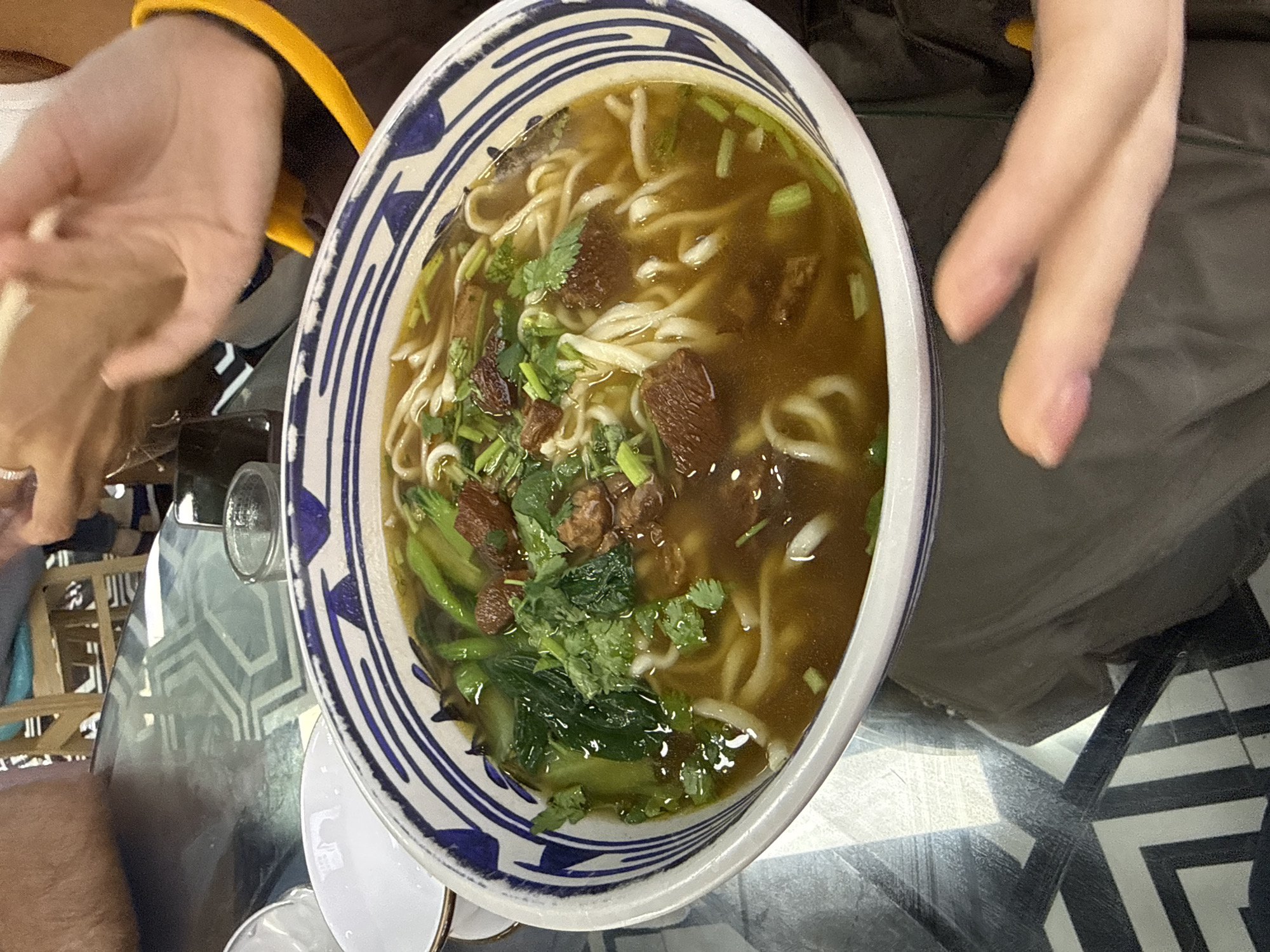 A high-angle shot depicts a bowl of beef noodle soup, held between two people's hands. The bowl is white with a blue patterned rim and contains noodles, green vegetables, cilantro, and chunks of beef in a broth. The setting appears to be a restaurant in the Dongcheng District, China, based on the style of the soup. The table is glass with a black and white geometric pattern underneath. A glass, a white saucer with a gold rim, and fragments of other furniture are also visible.