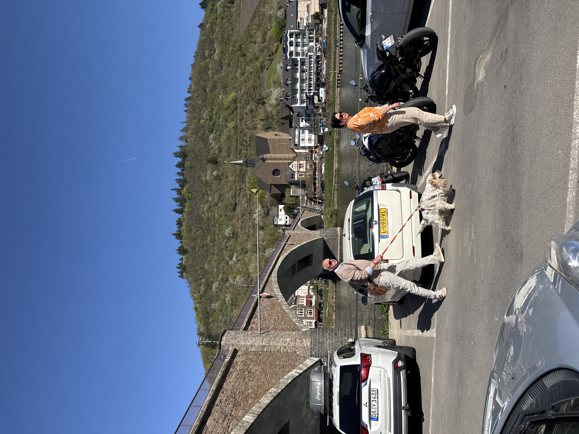 The image shows a sunny day in Cochem, Germany, along the Moselle River. Two men and a dog stand in a parking area. One man, in a light-colored suit and sunglasses, holds the dog's leash, while the other man, wearing an orange shirt, stands next to a parked motorcycle. A white van is parked nearby with a license plate starting with "L". A bridge and a church steeple are visible in the background, characteristic of Cochem's architecture. The Moselle River flows past the town, with buildings lining its banks. Cars and motorcycles are parked in the foreground. A hillside covered with trees rises in the background. The sky is clear blue with a faint contrail. The overall scene is a snapshot of everyday life in this picturesque town.