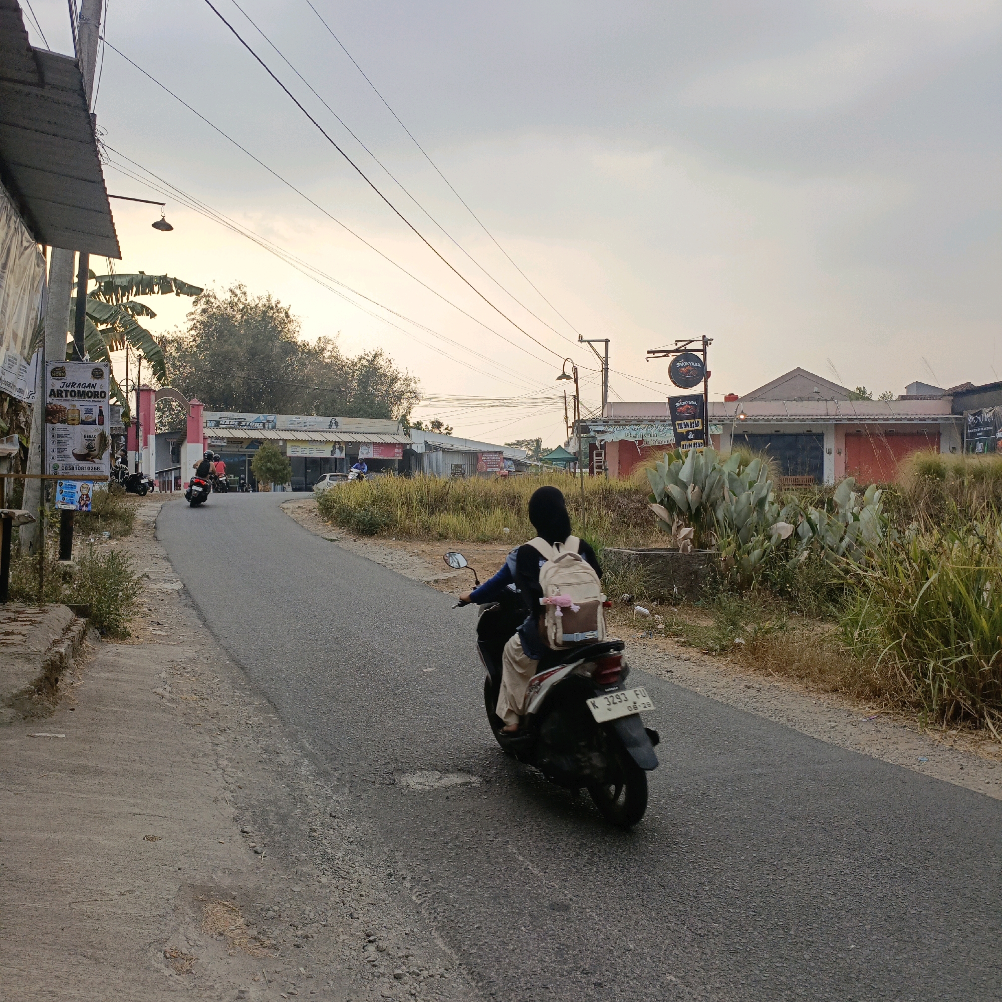 The image depicts a street scene in Jombor, Indonesia, during daylight hours. A person wearing a dark hijab and a light-colored backpack rides a dark-colored scooter on a paved road that curves to the left. The scooter's license plate is visible, reading "K 3293 FU". Buildings line the right side of the road, with some displaying signage, including one that reads "SMOKYARA". Tall, dry grass and vegetation grow along the roadside. Power lines crisscross the sky. On the left side of the road, a concrete sidewalk is visible, bordered by a curb. A pole with a street lamp and a sign that reads "JURAGAN ARTOMORO" is also present. In the background, more buildings and trees are visible, suggesting a semi-urban or suburban environment. Several other parked motorcycles are also present. The sky is overcast with a warm, soft light, indicating either early morning or late afternoon.