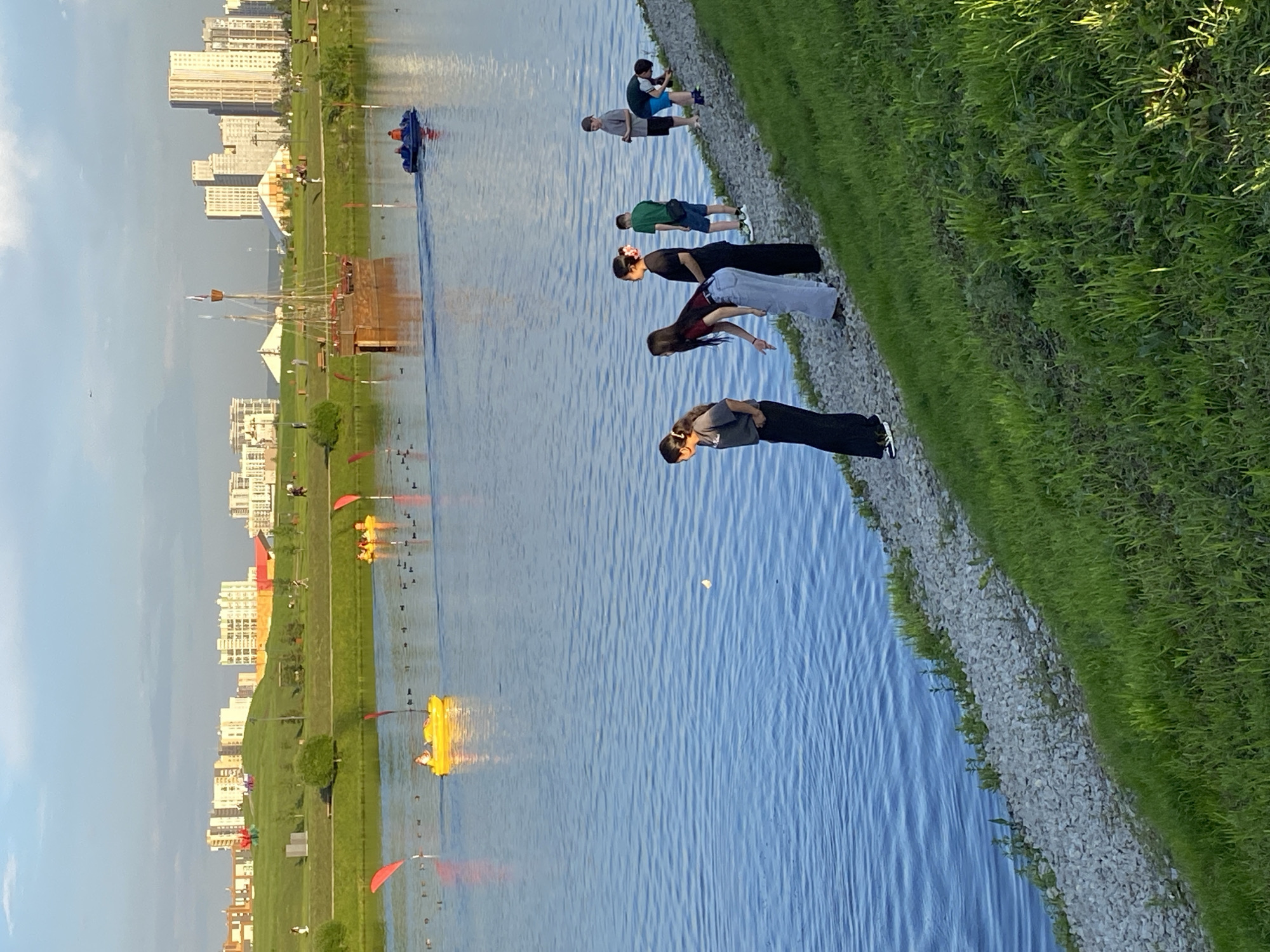 The image depicts the city of Yekaterinburg, Russia, specifically the pond at the city's zoo. The scene is set at sunset, with a golden light illuminating the water and the surrounding park. In the foreground, a group of people, mostly young, stand by the water's edge, some appearing to interact with the water. In the mid-ground, several yellow paddle boats are visible on the pond, with people enjoying leisure activities. A larger, replica wooden sailing ship is also present in the water, adding a historical element to the scene. The background shows a modern cityscape with tall buildings and green parkland, indicating a blend of urban development and natural spaces. The sky is a soft blue with hints of clouds, characteristic of a late afternoon or early evening.