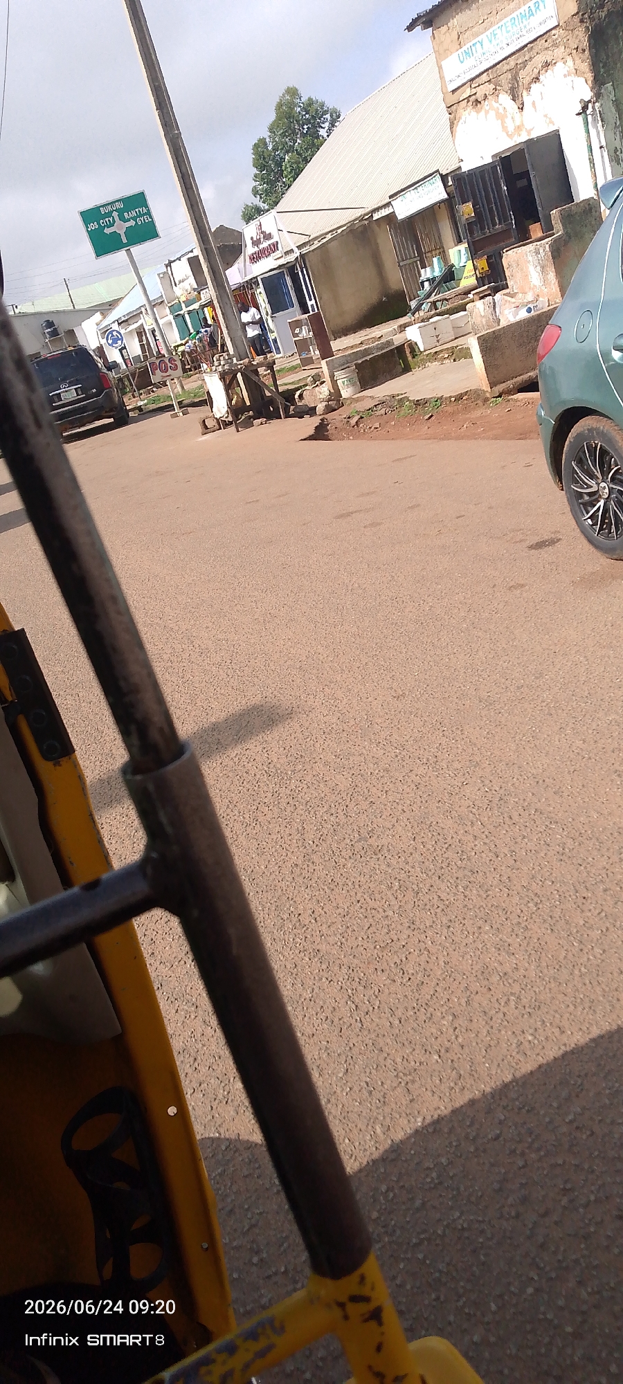 The image depicts a street scene in Jos, Nigeria. A green directional signpost indicates "BUKURU" to the left, "JOS CITY" to the straight ahead, and "RANTIYA-GYEL" to the right. Several businesses are visible along the street, including "UNITY VETERINARY" and a restaurant. A black SUV and a green car are parked on the roadside. In the foreground, part of a yellow motorized tricycle is visible, suggesting the photo was taken from within the vehicle. The road surface is asphalt, and the sky is overcast. The time stamp at the bottom indicates the photo was taken on June 24, 2026, at 09:20.