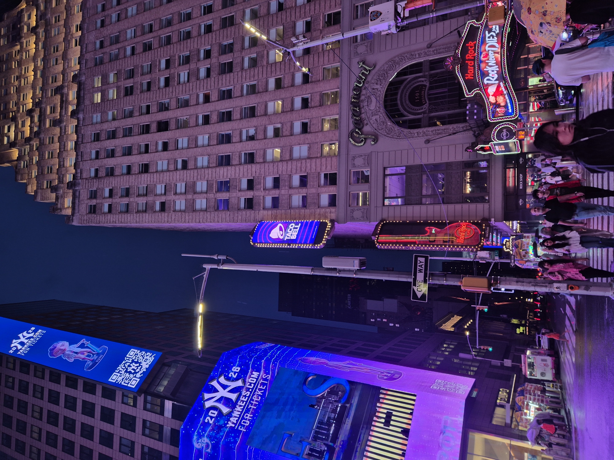 This is a low-angle, wide shot of Times Square in New York City at dusk. The scene is illuminated by a vibrant array of neon lights and digital billboards, casting a purple and blue glow over the street and surrounding buildings. The towering Paramount building, with its distinctive arched entrance, dominates the right side of the frame, its facade lit in a soft pink hue. Below, the marquee for the Hard Rock Cafe, with its "Rock Never Dies" slogan, glows in neon red and blue.
On the left, a massive digital billboard for the New York Yankees displays an advertisement with the team's logo and the text "YANKEES.COM FOR TICKETS." Another billboard above it shows a cartoon character and a QR code. The street is wet, suggesting recent rain, and a few pedestrians are visible walking on the sidewalk and crossing the street. A bus and various street vendors are also present. The sky above is a deep twilight blue.
In the foreground, several people are walking. A young woman in a black jacket is looking towards the camera. Behind her, a couple walks together, and further back, a man in a white t-shirt and another person holding a yellow umbrella are also visible. The overall impression is one of a bustling, dynamic urban environment at night.