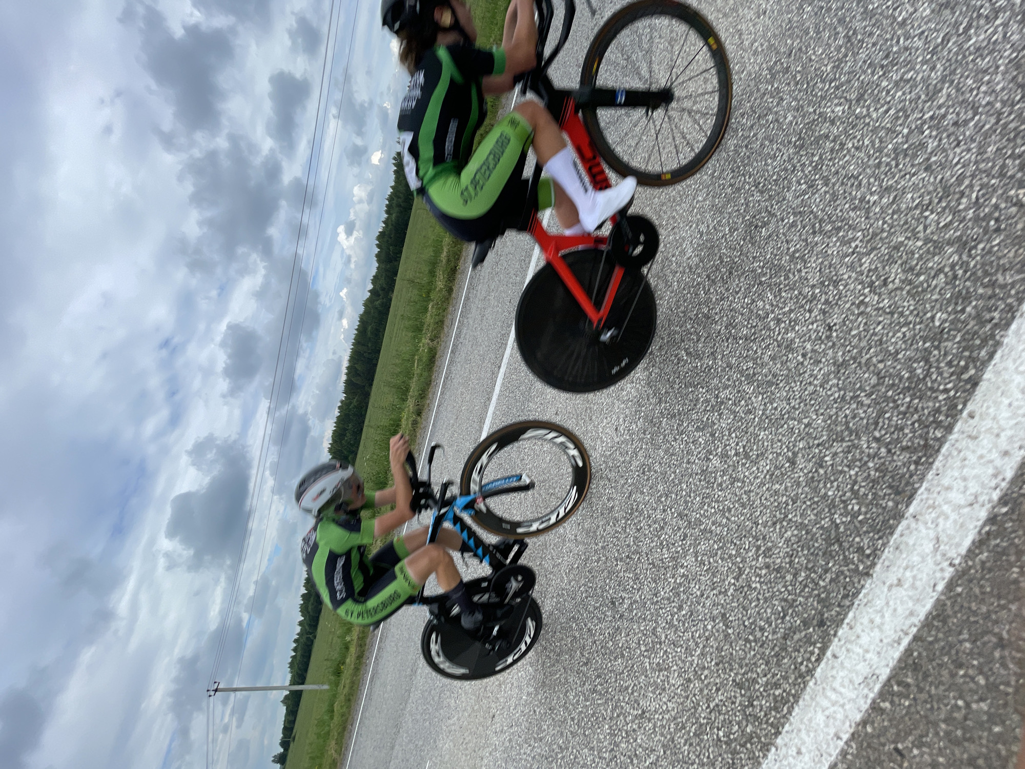 Two cyclists in green and black racing suits and helmets are riding on a paved road. They are both in a tucked position, leaning forward over their handlebars. The cyclist on the left is on a blue and black bicycle with disc wheels. The cyclist on the right is on a red and black bicycle, also with disc wheels. The road is grey asphalt with a white line on the left side. In the background, there is a field of green grass and a line of trees under a cloudy sky. The image is slightly blurry, indicating movement.