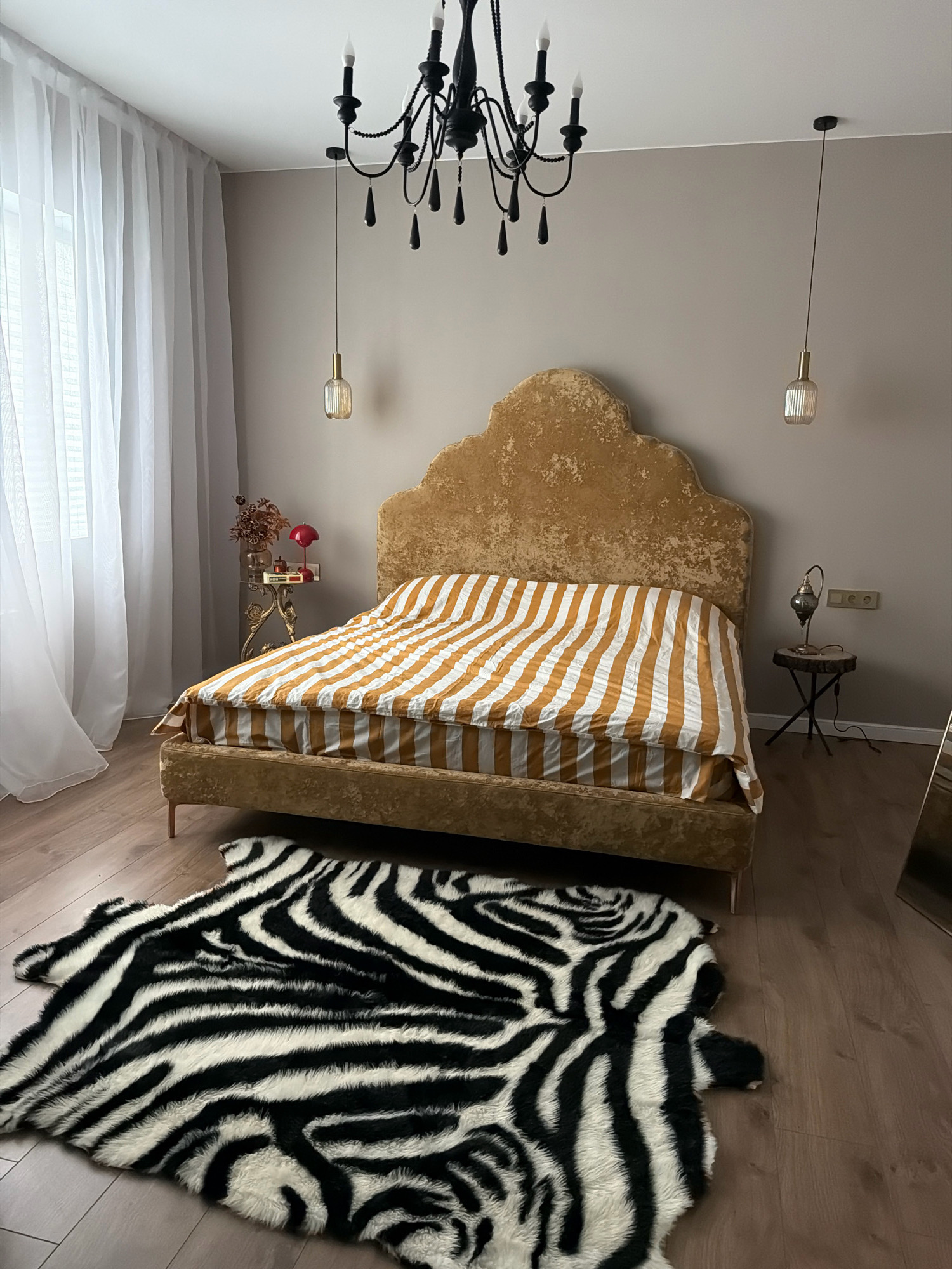This is a medium shot of a bedroom with a bed, a rug, and various decorative elements. The bed is gold-colored with a high, scalloped headboard and is covered with a duvet featuring a bold orange and white stripe pattern. In front of the bed, a faux zebra print rug is laid out on a light brown wooden floor. To the left of the bed, sheer white curtains hang over a window. A tall, ornate gold side table stands next to the left side of the bed, holding a vase with dried flowers and a red mushroom-shaped lamp. To the right of the bed, a small, rustic side table with a dark wood top and black metal legs supports a decorative lamp. A black chandelier hangs from the ceiling, with two additional modern pendant lights on either side. The walls are painted a neutral taupe color. The overall impression is one of eclectic and dramatic decor. The image does not contain any information that would allow for the determination of its location in the city of Orsha, Belarus.