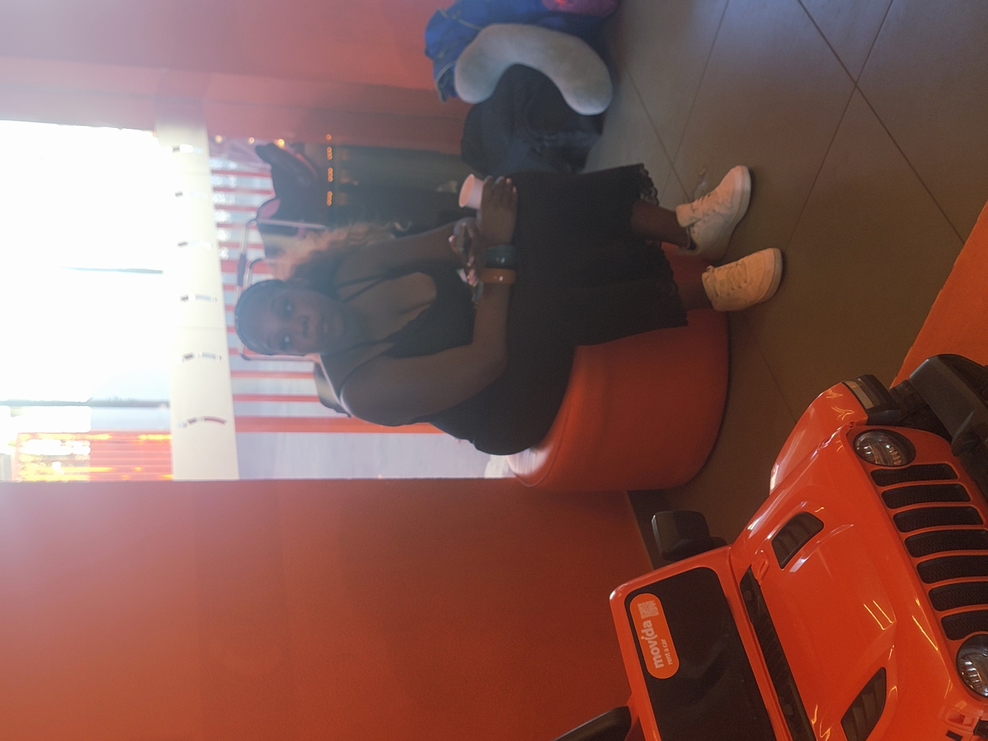 A Black woman is seated on an orange ottoman in a brightly lit indoor space, possibly a play area or waiting room. She is wearing a black dress with lace trim and white sneakers. Her hands are clasped in her lap, and she holds a white cup. Behind her, there are bags and a large, light-colored pillow. To her left, the front of a bright orange toy car is visible, with a "movida rent a car" sticker on it. The background features an orange wall and large windows with vertical blinds, suggesting a location in Faro, Portugal.