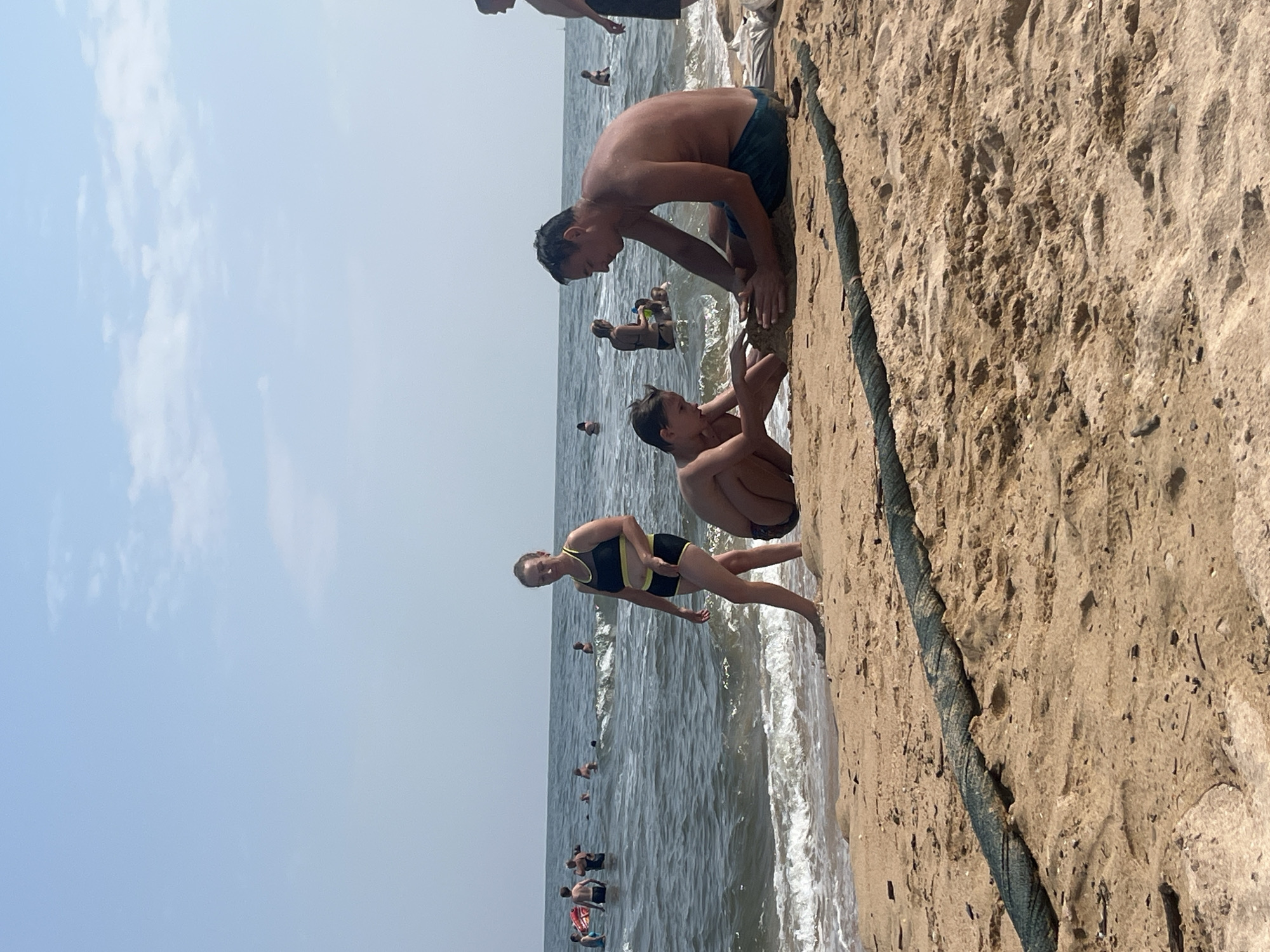The image depicts a beach scene in Golubitskaya, Russia, with several people engaged in various activities. In the foreground, a man and a boy are sitting on the sand, appearing to build a sandcastle. The man is shirtless and wearing blue swim trunks, while the boy is also shirtless. To the left, a woman in a black and yellow swimsuit stands near the water's edge, looking to her left. In the background, numerous other individuals are visible in the sea, some swimming and others standing in the shallow water. The sea is calm with small waves reaching the shore, and the sky is clear with scattered clouds. A thick, dark green rope lies on the sand in the foreground, stretching across the image.