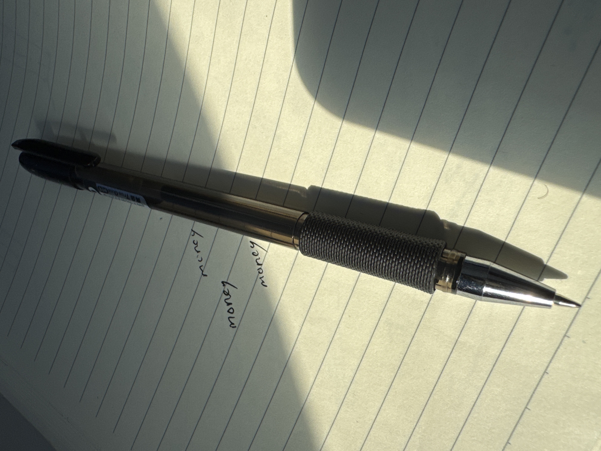 A pen lies on a lined notebook. The word "money" is written three times on the notebook page. The pen has a black cap and a metallic tip with a textured grip. Sunlight casts a shadow across the notebook. There is no information about the location in Guta, China.