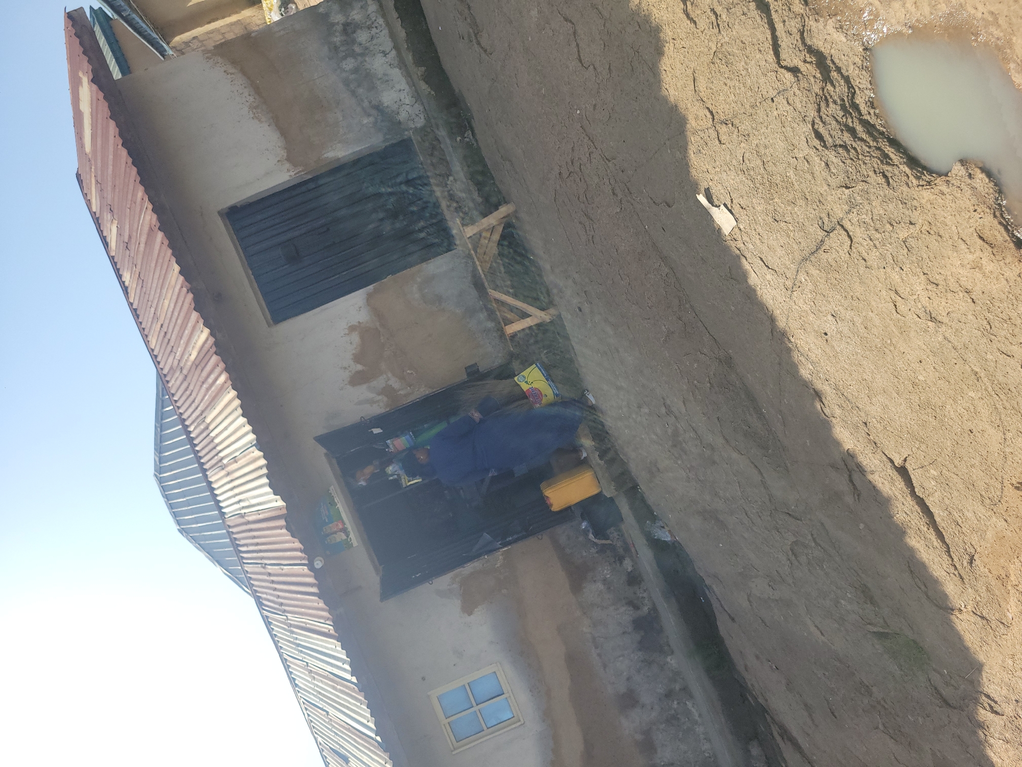 A person in a blue robe stands in the doorway of a small, open-air shop in Jos, Nigeria. The shop has a corrugated metal roof and concrete walls. A yellow jerrycan sits to the left of the doorway, and a wooden bench is placed in front of the shop. The ground in front of the shop is dirt and cracked, with a small puddle of water to the right. The sky is bright and clear.