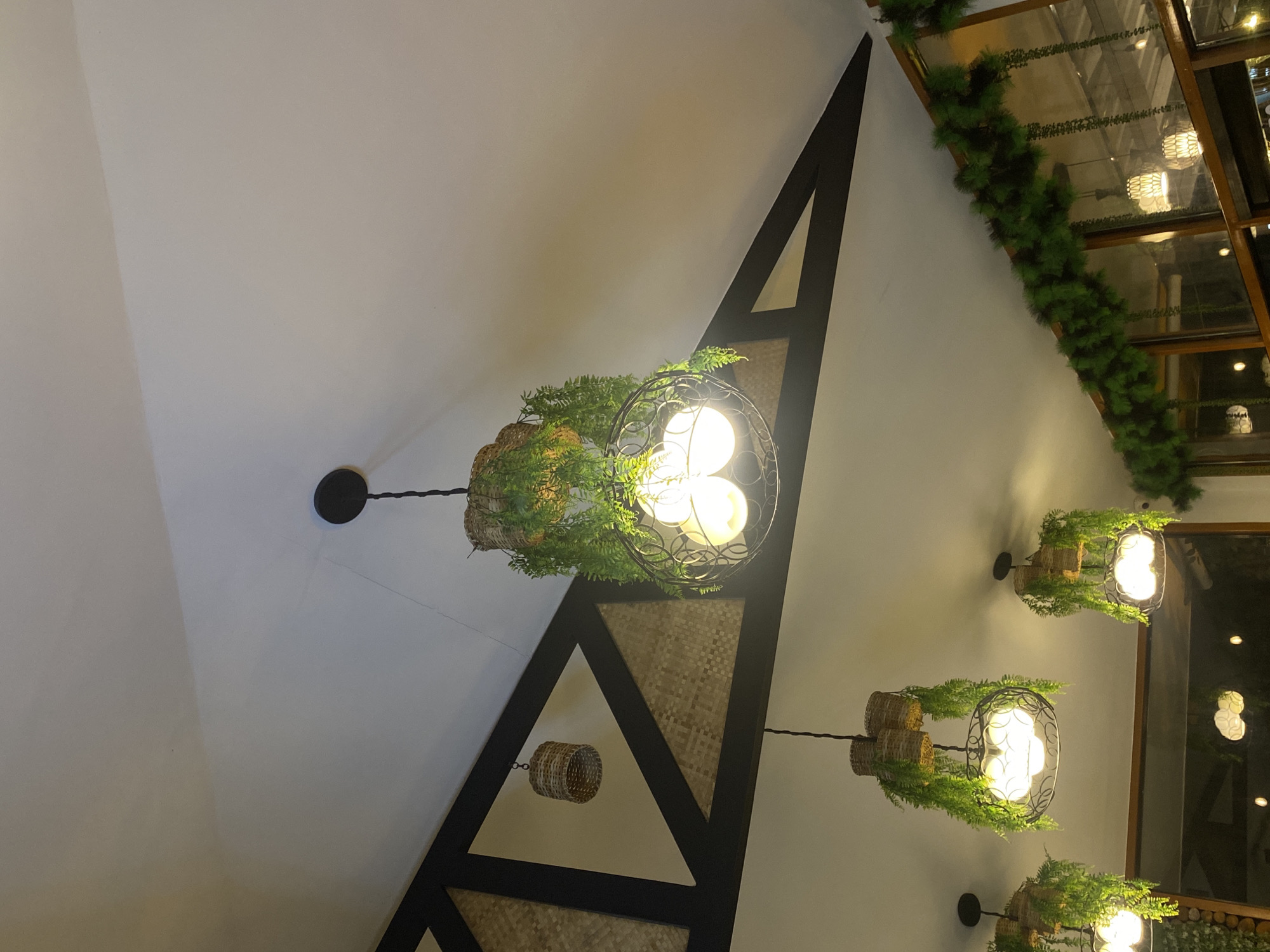 The image shows a ceiling in Davao City, Philippines, decorated with hanging light fixtures and greenery. Three main pendant lights are visible, each featuring a woven basket-like structure at the top adorned with faux green foliage. Below the baskets, the lights themselves are encased in decorative metal cages and emit a warm, yellow glow from what appear to be multiple bulbs. A smaller woven basket hangs from the triangular beam structure on the left. Along the top of a window or glass partition in the background, a garland of artificial green pine branches is draped. The ceiling is white, and dark beams create a geometric pattern. The overall ambiance suggests an indoor setting, possibly a restaurant or cafe, with a rustic and naturalistic aesthetic.