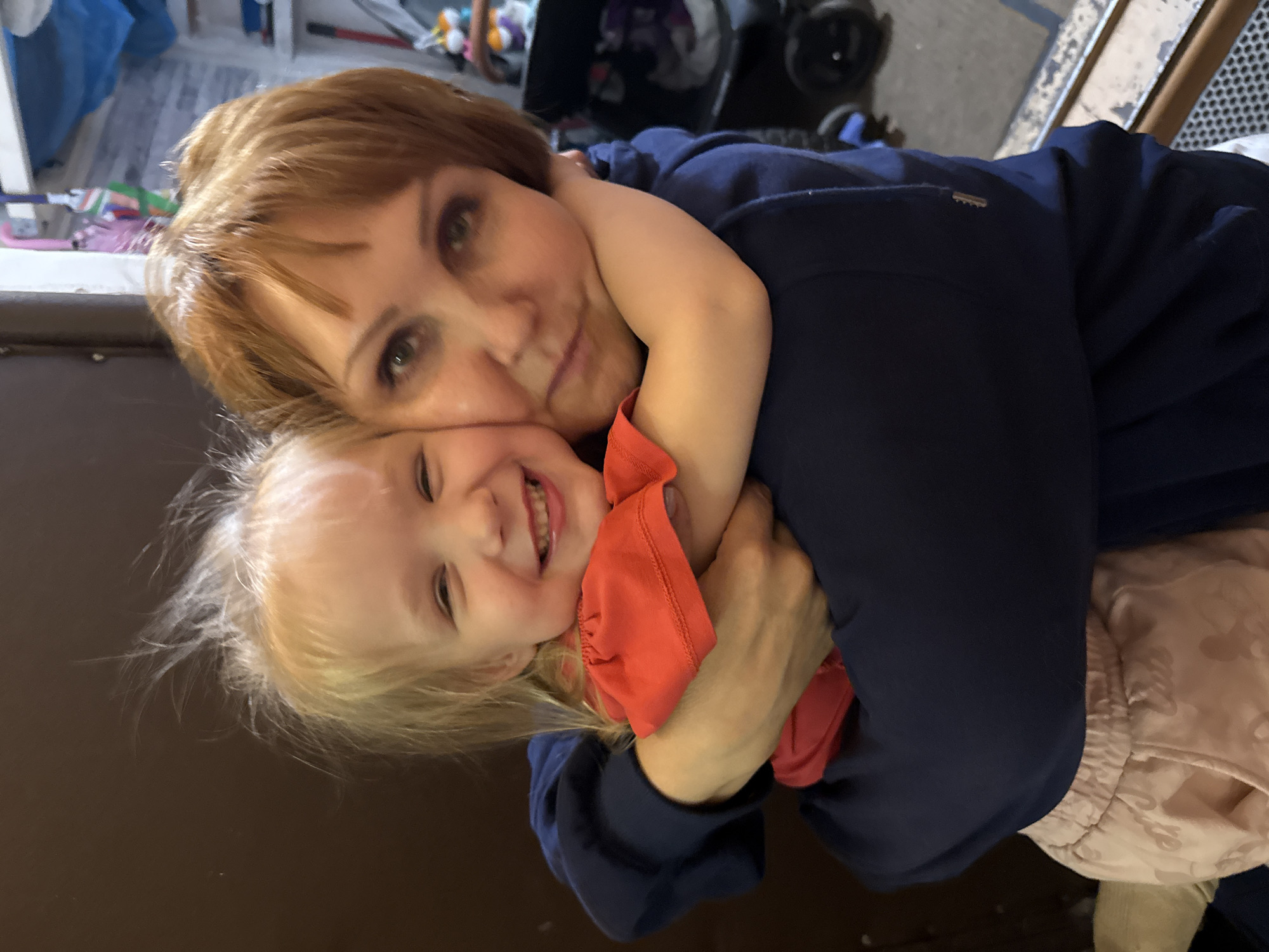 An adult woman with short auburn hair and eye makeup embraces a young child with blonde hair. The woman is wearing a dark blue hoodie, and the child is wearing a bright red top and light pink patterned pants. They are holding each other tightly, with the child's arms around the woman's neck and the woman's arms wrapped around the child. Both appear to be smiling. The background is blurred but suggests an indoor setting with a dark brown wall on the left and a cluttered area with shelves and objects on the right.