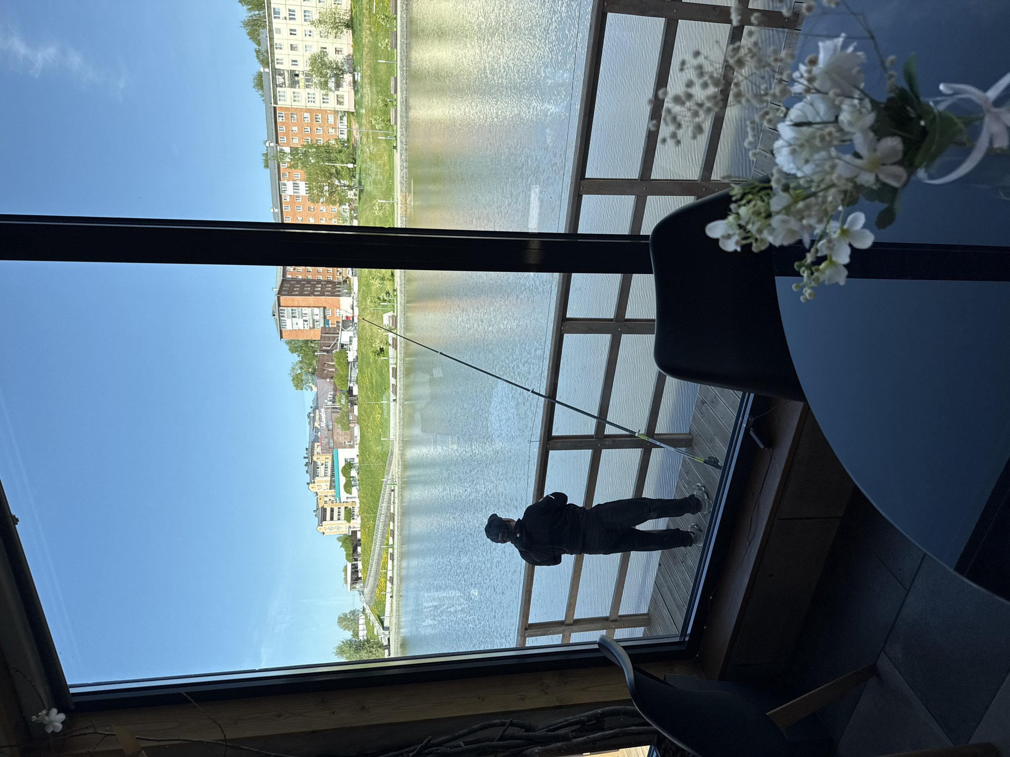From the perspective of inside a building with large windows, the image depicts a view across a body of water towards a city landscape. A man is standing outside, fishing off a wooden structure next to the water. Buildings of varying heights and colors, typical of urban architecture, are visible in the background. The sky is clear and blue. A table and floral arrangement are partially visible inside the building, indicating an indoor space. The city is presumed to be Votkinsk, Russia.