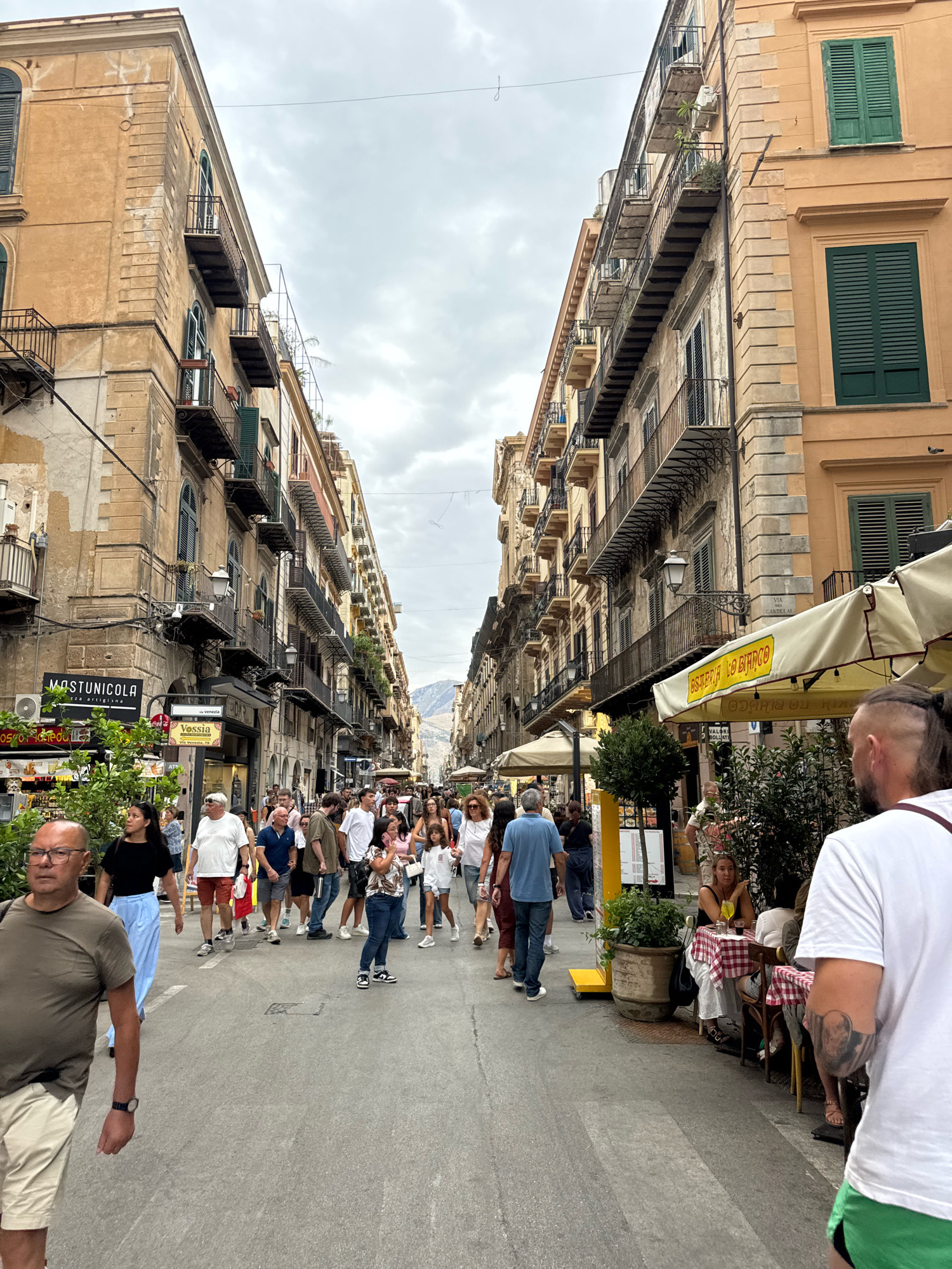 The image shows a bustling street scene in Palermo, Italy. The street is lined with tall, old buildings featuring balconies and shuttered windows. People are walking in both directions, some are dining at outdoor tables of a cafe. The overall impression is of a lively, urban environment on an overcast day.