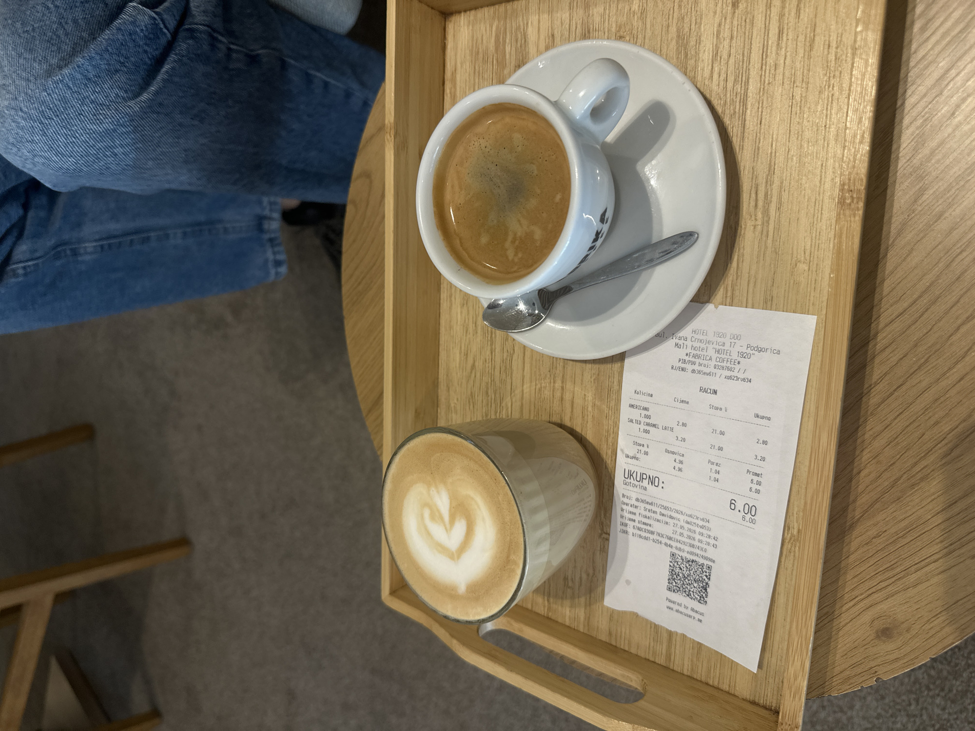 A wooden tray sits on a table. On the tray are an espresso in a white cup and saucer with a silver spoon, a latte in a clear glass, and a receipt. The receipt lists the items as "Americano" and "Salted Caramel Latte" from the establishment "FABRICA COFFEE" at "Hotel 1920", located at "Ivana Crnojevica 17" in Podgorica, Montenegro. A person wearing blue jeans is partially visible to the left of the tray. The edge of a wooden chair is visible in the lower left.