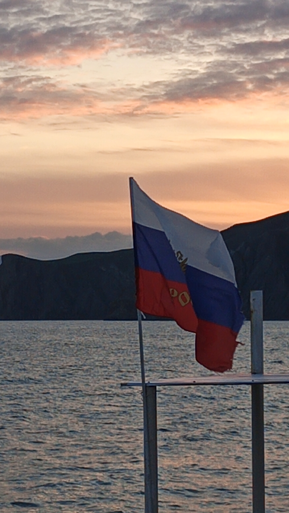 A Russian Navy flag is seen waving in the wind against a backdrop of water, land, and a sky illuminated by a sunset. The flag is mounted on a pole structure that is partially visible. The water is calm, and the land appears to be a mountainous area. The sky is filled with clouds and the light from the setting sun. The location is Unknown, Unknown.