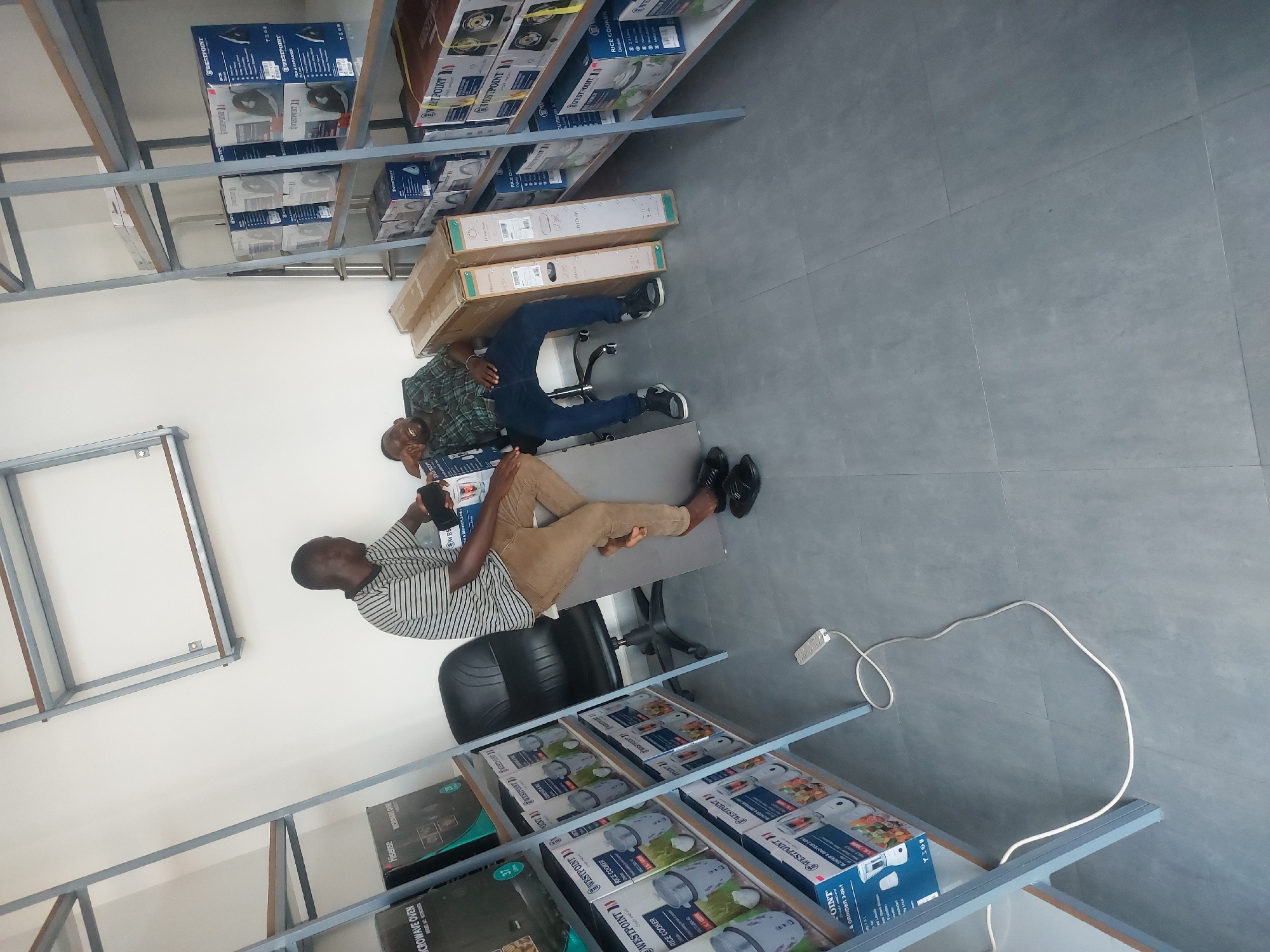 Two African men are in a store that appears to sell house appliances such as microwave ovens and rice cookers. The man closest to the camera is wearing a striped shirt and tan pants. He is sitting on a black office chair, looking down at his phone. The second man is wearing a plaid shirt and jeans and is reclined on top of two large boxes, using them as a seat. The products on the shelves have the brand name Westpoint. The exact location of this store in the city of Dar es-Salaam, Tanzania is not apparent from the image.