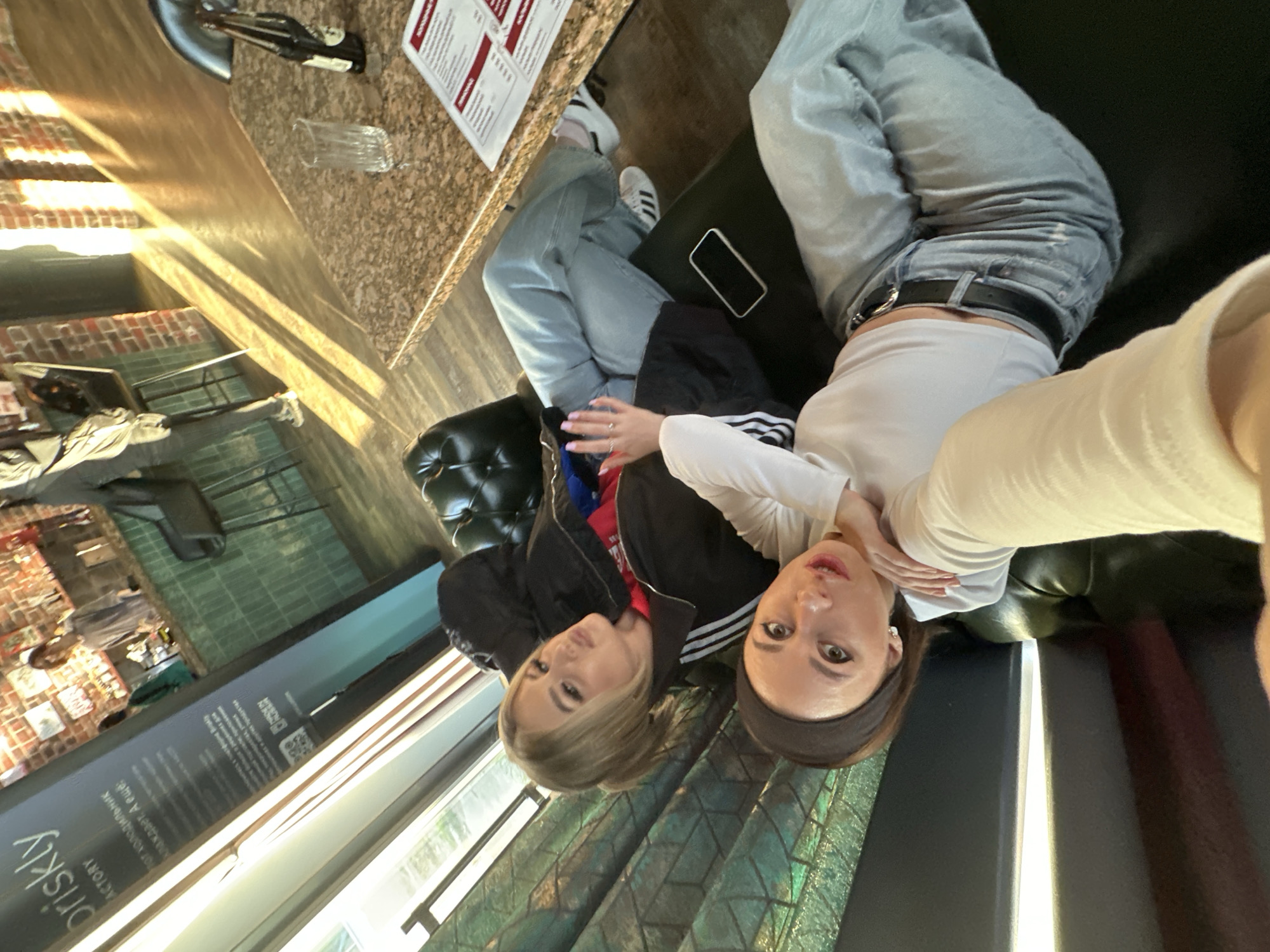 Two women are posing for a selfie inside a restaurant. One has blonde hair and is wearing a black jacket with Adidas stripes and light blue jeans, and the other woman is wearing a white top and light blue jeans with a belt. There is a table next to them with a menu, a glass, and what appears to be a bottle of hot sauce. The scene is in a restaurant in Votkinsk, Russia. The restaurant has a window overlooking what seems to be an outdoor area with plants.