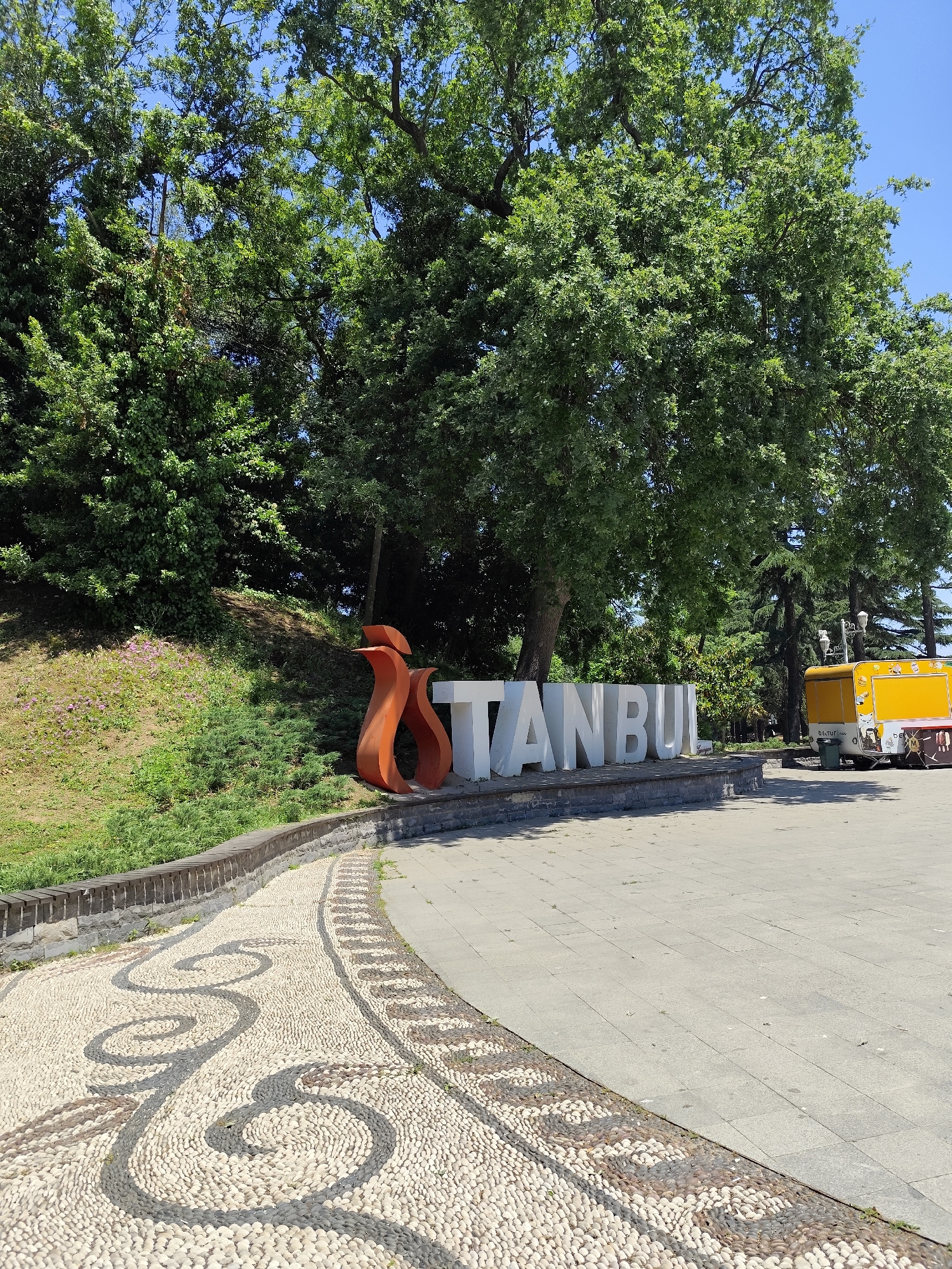 The image captures an outdoor scene in what appears to be a public park or plaza in Sarıyer, Turkey, under a clear blue sky on a bright, sunny day. The prominent feature is a large, white block letter sign spelling "ISTANBUL". The initial letter 'I' is replaced by an artistic, flame-like or tulip-shaped sculpture in a vibrant orange hue.
To the left of the sign, a grassy slope rises, covered with green foliage and hints of small purple flowers. This incline is bordered by a low stone wall topped with wooden planks, alongside which runs a decorative pebbled path. This path features intricate swirling patterns created with darker pebbles against a lighter stone base, leading towards the foreground.
The ground in the mid-to-right section of the image is a spacious paved area, likely a pedestrian walkway or square. Further to the right and slightly behind the sign, a bright yellow food truck is visible, parked on this paved surface. The truck has various cartoon illustrations on its side and some indistinct text, possibly "b.stur".
The background is dominated by lush, dense green trees, suggesting a natural and verdant environment. The sunlight creates distinct shadows, indicating a high sun position. There are no people visible, and no specific event or activity is taking place, presenting a tranquil and inviting public space.