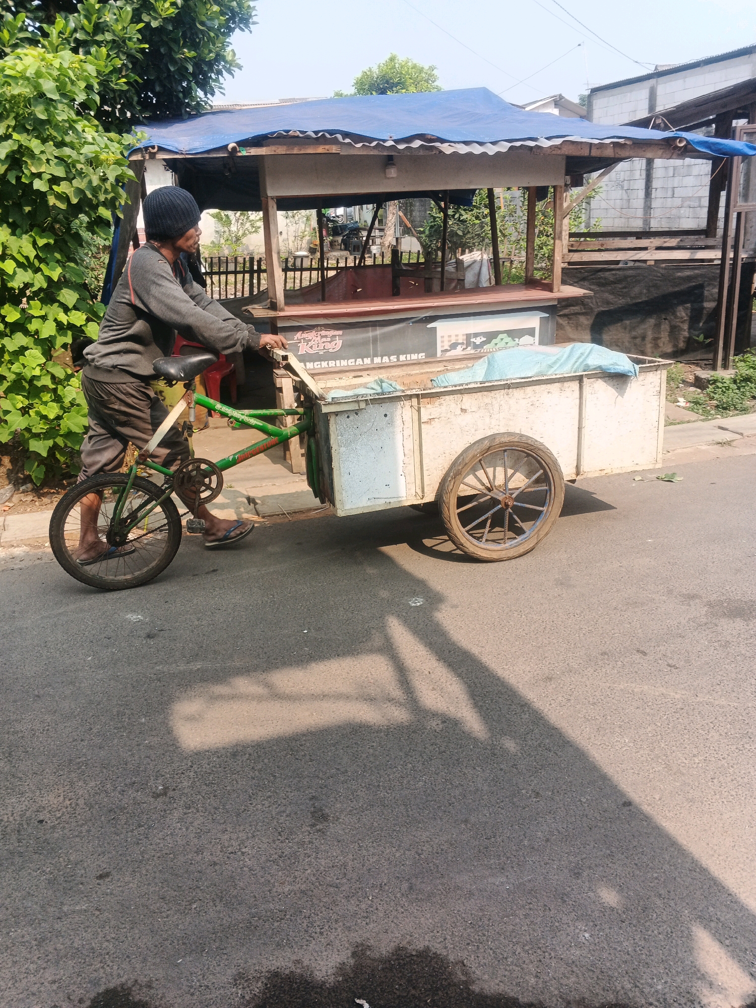 In Kadu, Indonesia, a person wearing a dark beanie, gray long-sleeved shirt, and dark pants is pushing a modified bicycle with a large cargo trailer attached. The bicycle has a single rear wheel and a front wheel that supports the trailer. The trailer is constructed of white panels and has a light blue tarp covering part of its contents. The person is pushing the trailer from behind, with their hands on the trailer's frame. The scene takes place on an asphalt road, with a green leafy tree on the left and a makeshift stall with a blue tarp roof in the background. The stall appears to be made of wood and corrugated metal. The sky is clear and bright.