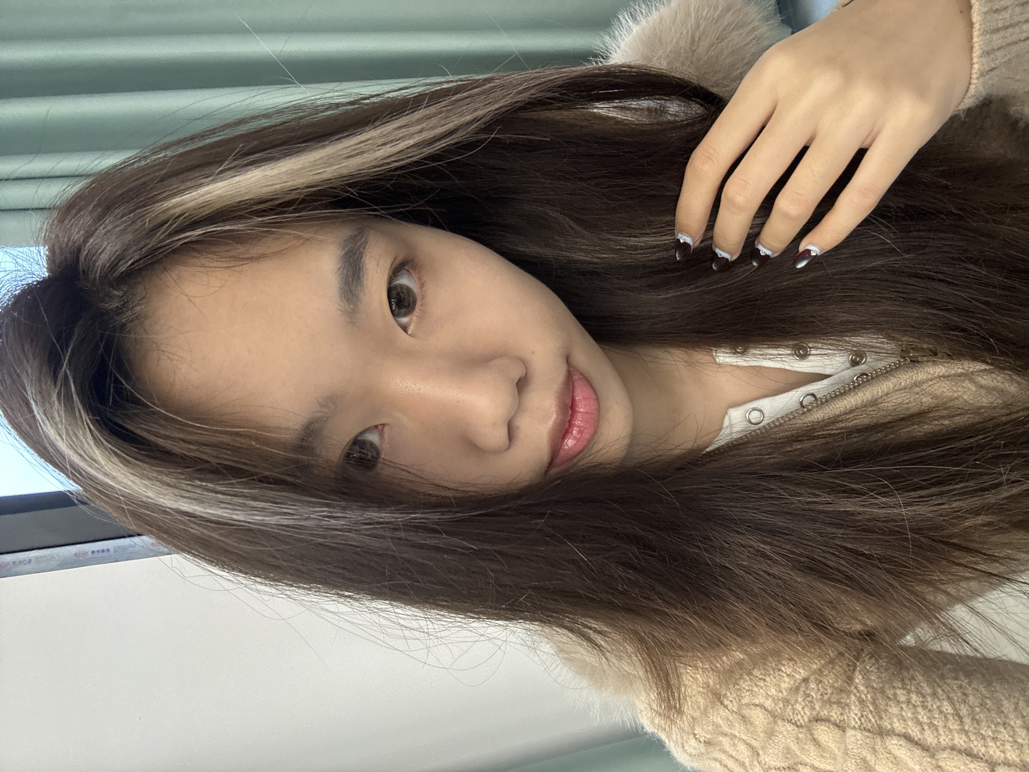 This image depicts a close-up, chest-up selfie of a single East Asian female subject, located indoors within Nan'an District, China. The subject has long, dark brown hair with distinct light blonde or silver highlights framing the right side of her face. Her eyes are brown, and her expression is neutral as she looks directly at the camera. She is wearing a light beige or brown cable-knit cardigan featuring a fuzzy collar or trim, layered over a white top with a visible zippered or buttoned placket. Her left hand, adorned with dark-colored nail polish featuring white or light blue tips and small decorative elements, is resting on her hair near her shoulder. The background consists of a plain white wall or surface on the left and partially visible teal or light green vertical blinds or curtains on the right, suggesting a room with a window.