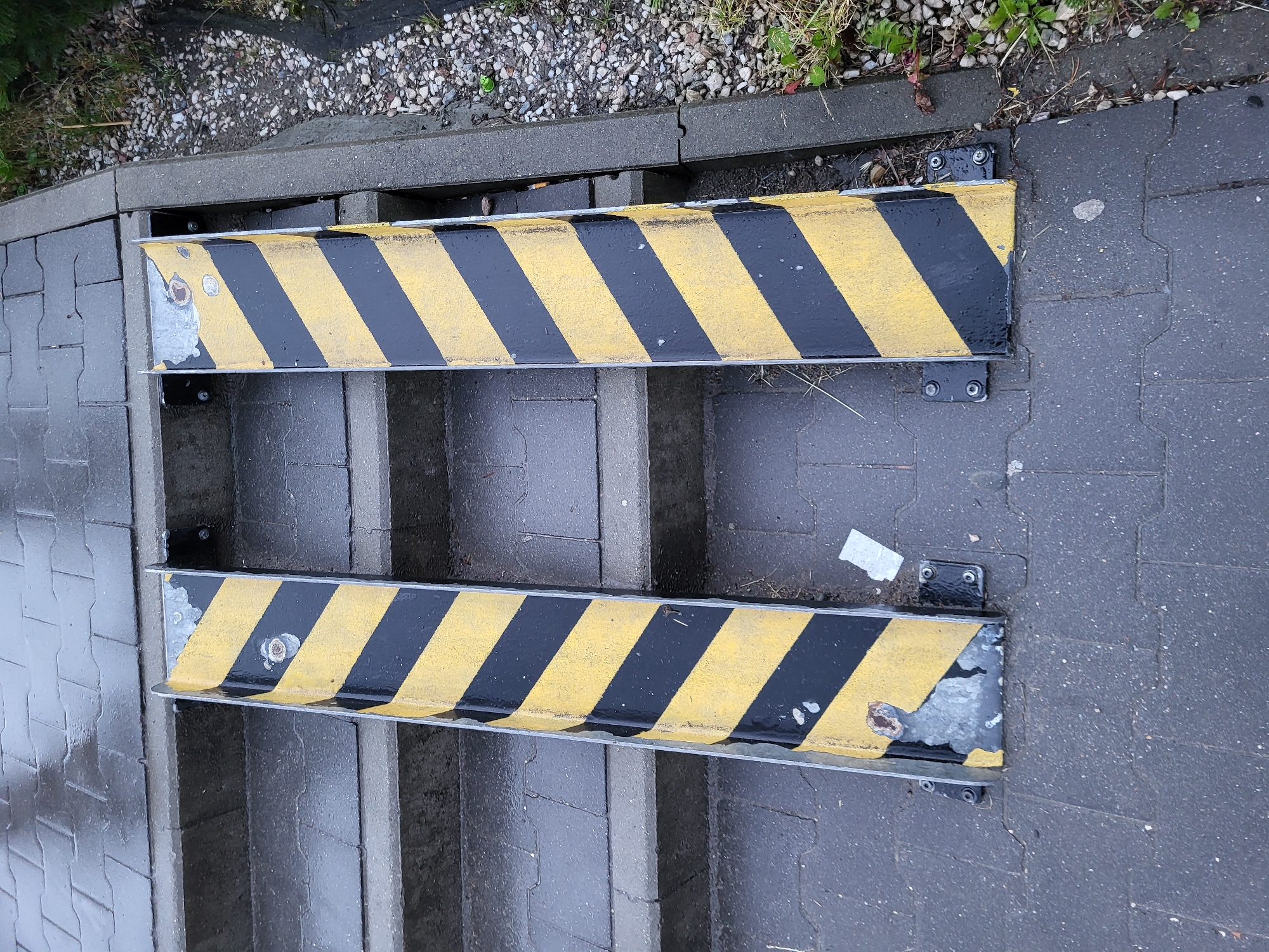 The image presents an overhead view of an outdoor pathway featuring two parallel metal barriers with distinctive black and yellow diagonal stripes. These barriers appear to serve as ramps or guides, likely intended for rolling items down steps or an incline. They are mounted above grey concrete steps or slabs.
To the left of the barriers, the ground is paved with interlocking grey bricks, while to the right, it transitions to a darker, possibly asphalt or textured concrete surface. At the top of the frame, there is a section of gravel and small stones with sparse green vegetation, suggesting a garden edge or natural border.
No specific activity is taking place; the scene is static. The surfaces of the pavement, steps, and barriers appear wet, indicating recent rain or damp conditions. A notable detail is the worn condition of the yellow paint on the barriers, which is chipped and faded in various spots, revealing the underlying metal that shows signs of rust and corrosion, particularly near the attachment points. The metal plates and bolts securing the barriers are clearly visible. The lighting suggests it is daytime, possibly overcast due to the soft, diffused illumination and wet surfaces. No discernable text is visible anywhere in the image.
