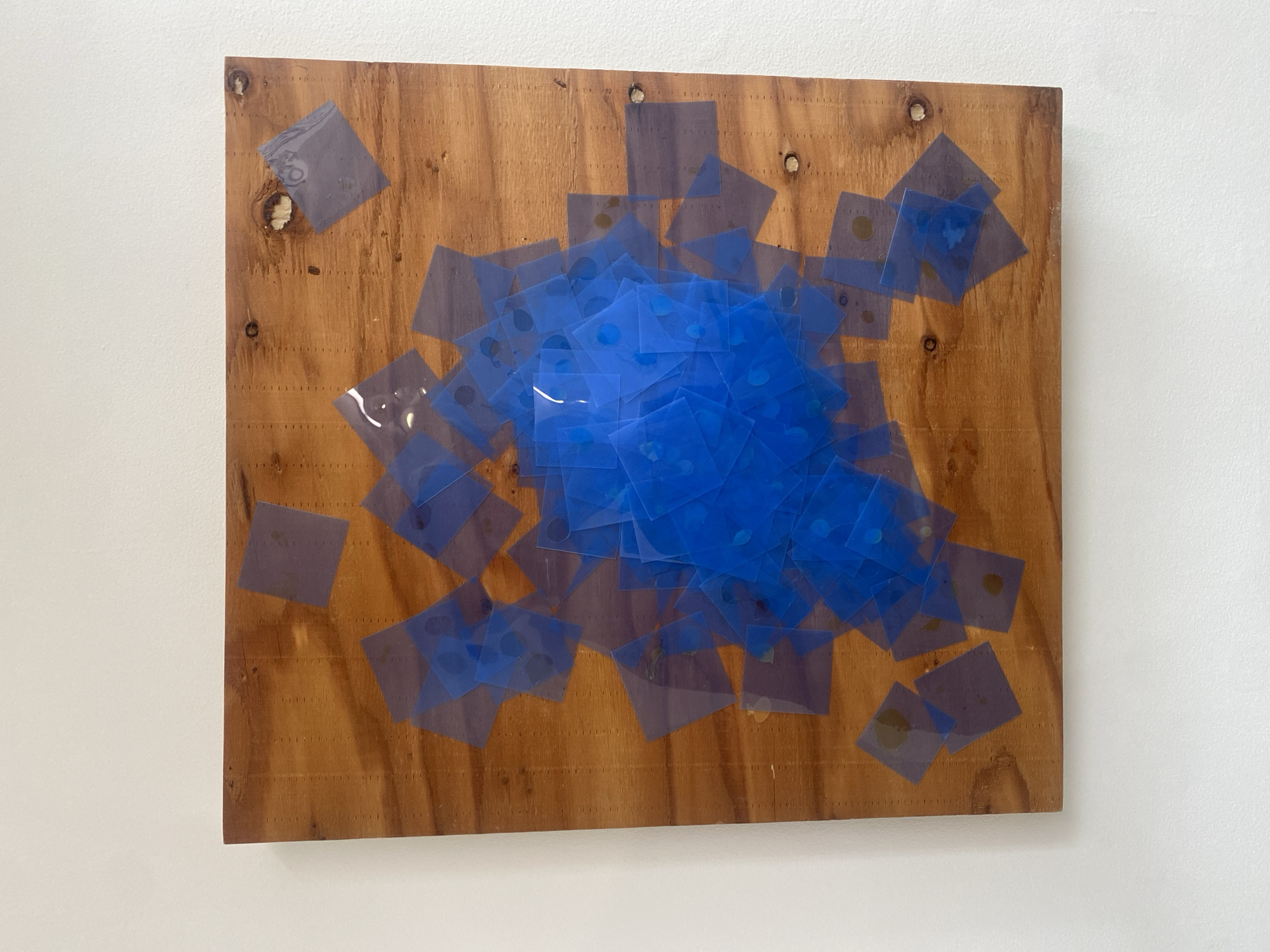 An artwork or decorative panel is displayed on a plain, off-white wall. The central subject is a rectangular piece of light to medium-brown wood, possibly plywood, with a visible wood grain and several knot holes. The wooden panel features numerous small, regularly spaced vertical lines of indentations across its surface, suggesting a grid or pegboard-like structure, though no pegs are present. In the upper part of the panel, some circular indentations or holes are visible, with what appear to be silver-colored nails or screws securing the panel to the wall.
Overlaid on the wooden panel is a dense, irregular cluster of small, square pieces of translucent blue material. These squares are layered over each other, creating varying shades of blue and depth, with the most concentrated areas appearing as a vibrant, solid blue. Individual blue squares extend outward from the central mass, scattering across the wooden background. Beneath some of the blue squares, small, dark, circular or irregular markings are visible, possibly trapped air, adhesive residue, or intrinsic design elements.
The artwork is presented against a neutral, brightly lit background, typical of an indoor setting such as a gallery, home, or studio. No people or specific activities are depicted. The overall impression is that of an abstract or mixed-media art piece. No discernible text is visible on the artwork or the surrounding wall. The location context for this display is Paris, France.