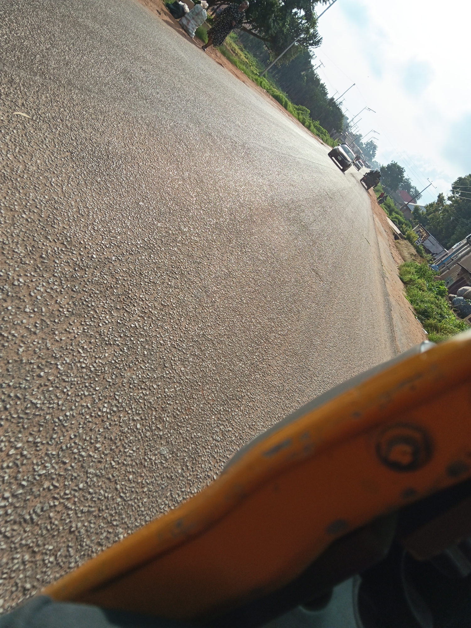 The image depicts a sun-drenched, asphalt road in Vom, Nigeria, with a slight upward tilt to the camera angle. The road surface is a textured grey asphalt, sparsely populated with small, light-colored stones, creating a speckled appearance. On the left side of the road, blurred figures of people are visible near stacked items, possibly merchandise or building materials. Further down the road, a white car drives away from the camera, followed by another vehicle. To the right of the road, a strip of green grass borders the asphalt, and beyond that, trees and a few buildings are present. In the foreground, the edge of a bright yellow object, likely a vehicle part, is visible, partially obscuring the view of the road. The sky is a bright, cloudy white.