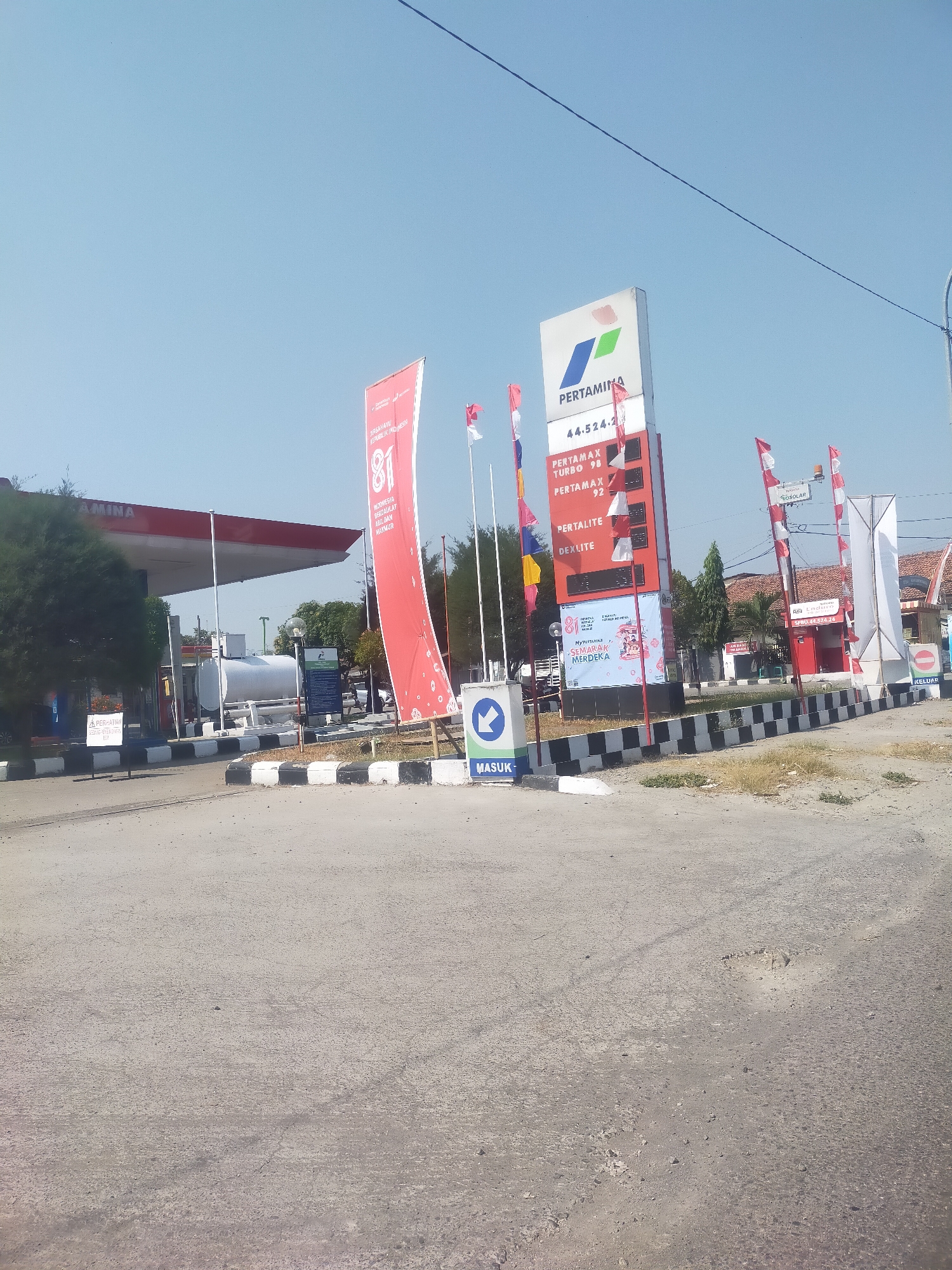 A street-level, eye-level, medium shot captures a Pertamina gas station under a clear blue sky in Kalikangkung, Indonesia. The gas station features a prominent red and white canopy extending over multiple fuel pumps. A large red banner with the text "INDONESIA SEMARAK MERDEKA" is displayed vertically, adorned with the number "81".
To the right of the canopy, a tall, white sign displays the Pertamina logo and fuel prices. Visible prices include "PERTAMAX TURBO 98", "PERTAMAX 92", "PERTALITE", and "DEXLITE". Several smaller red and white flags on poles are positioned in front of the fuel price sign. A blue and white directional sign with a left-pointing arrow and the word "MASUK" (meaning "Enter") is visible to the left of the fuel price sign.
A white storage tank is situated behind the fuel pumps, to the left of the red banner. A black and white checkered border runs along the edge of the paved area. The ground in the foreground is asphalt, with some visible cracks and potholes. In the background, trees and buildings are visible, suggesting an urban or suburban environment. No people are clearly visible in the frame.