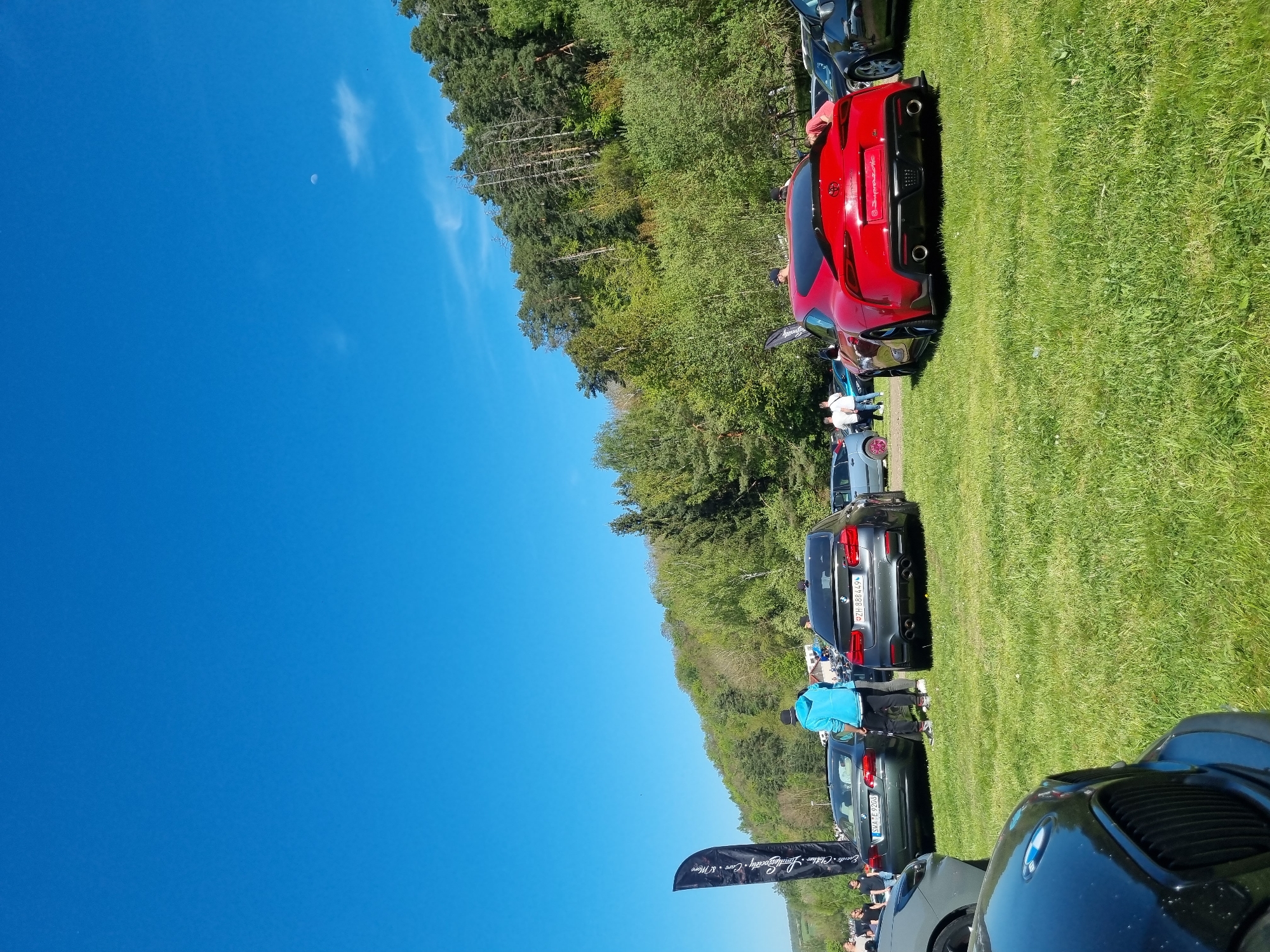 The image shows a car meet on a grassy field, likely in Geiselwind, Germany, given the license plates. Several cars are parked in a line, including a red Toyota Supra, a black BMW M5, and other BMW models. People are seen standing near the cars, possibly interacting and observing the vehicles. In the background, there is a dense forest under a clear blue sky. A black banner with white text and a logo is visible near the foreground.
