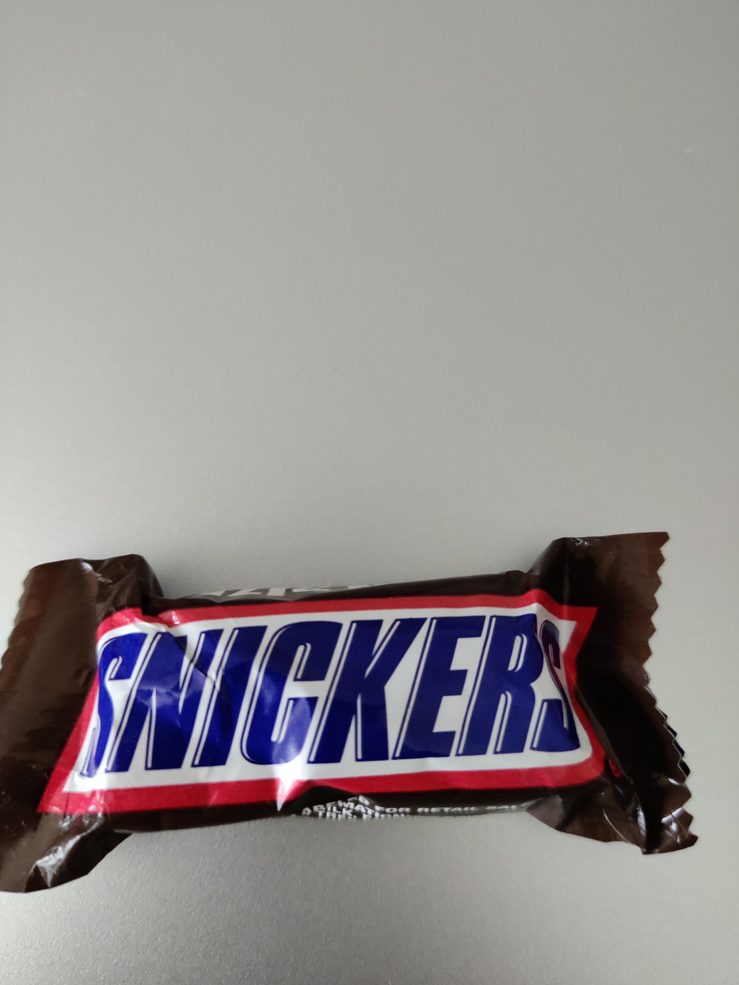 A Snickers candy bar wrapper is centered horizontally and positioned towards the bottom of the frame. The wrapper is predominantly brown with the brand name "SNICKERS" emblazoned in large blue letters with white outlines, set against a red and white stripe. The background is a plain, light-colored, smooth surface, likely a wall or tabletop, with a subtle shadow cast by the candy bar wrapper. The image does not provide any information about the specific location within Lower Paxton Township, United States of America.