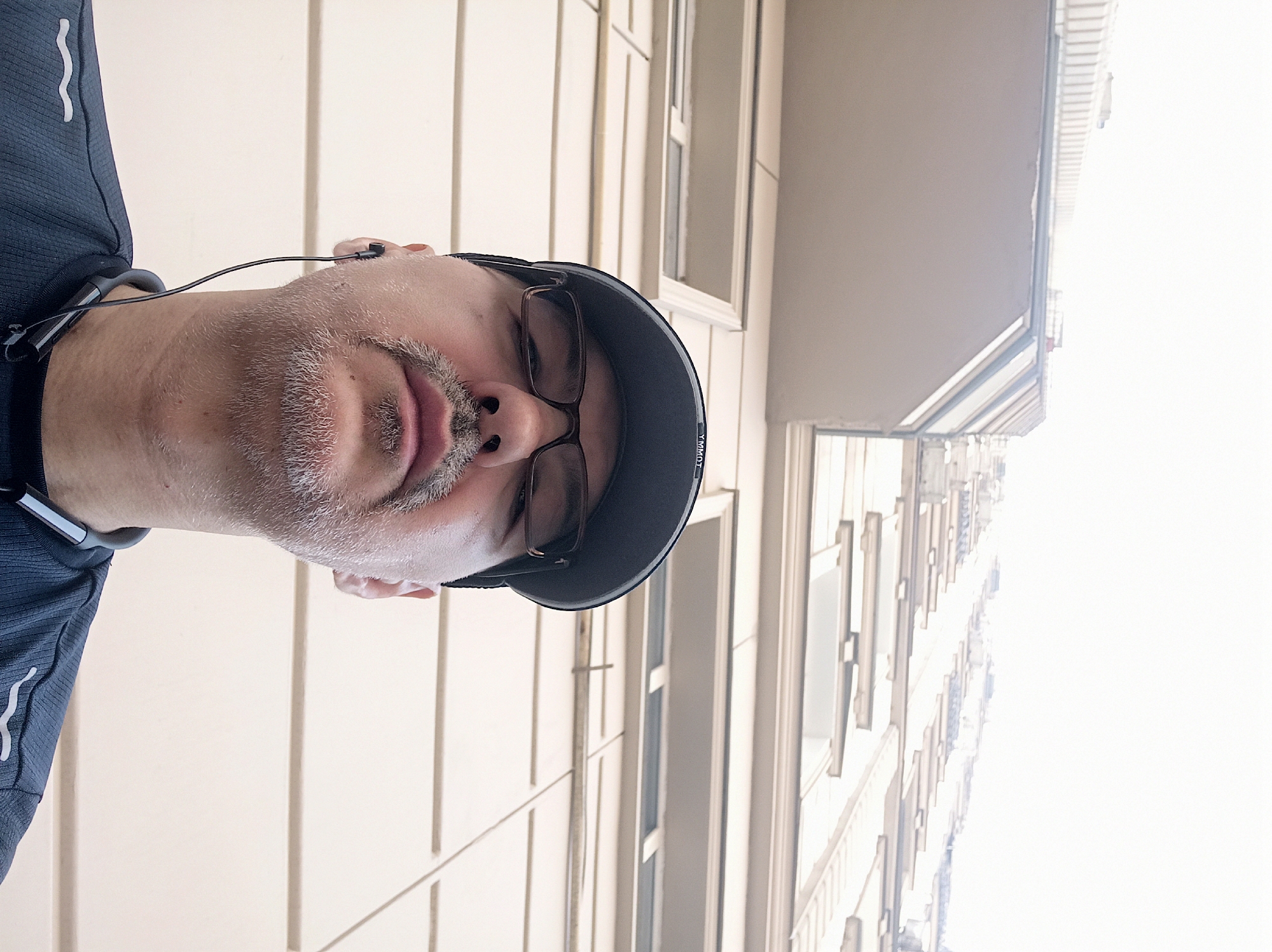 The image is a low-angle, wide-shot selfie taken outdoors. The subject, a man with a goatee and wearing glasses and a baseball cap, is looking upwards. He is wearing a dark blue athletic shirt with white accents and earbuds. The background is a tall, beige building with multiple windows and balconies, shot from below looking upwards towards the sky. The building's architecture suggests it is a modern apartment or office building. The sky appears overcast and bright, indicating it is daytime. This image was taken in Dushanbe, Tajikistan.